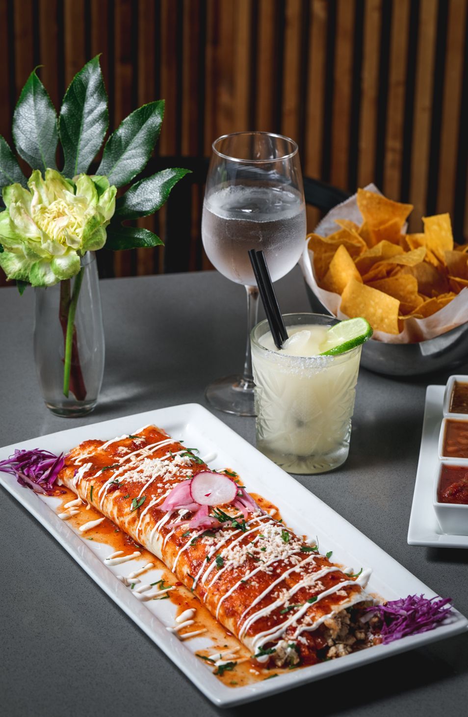 Burrito on a white plate, margarita, water glass, chips, and salsa on a gray table.