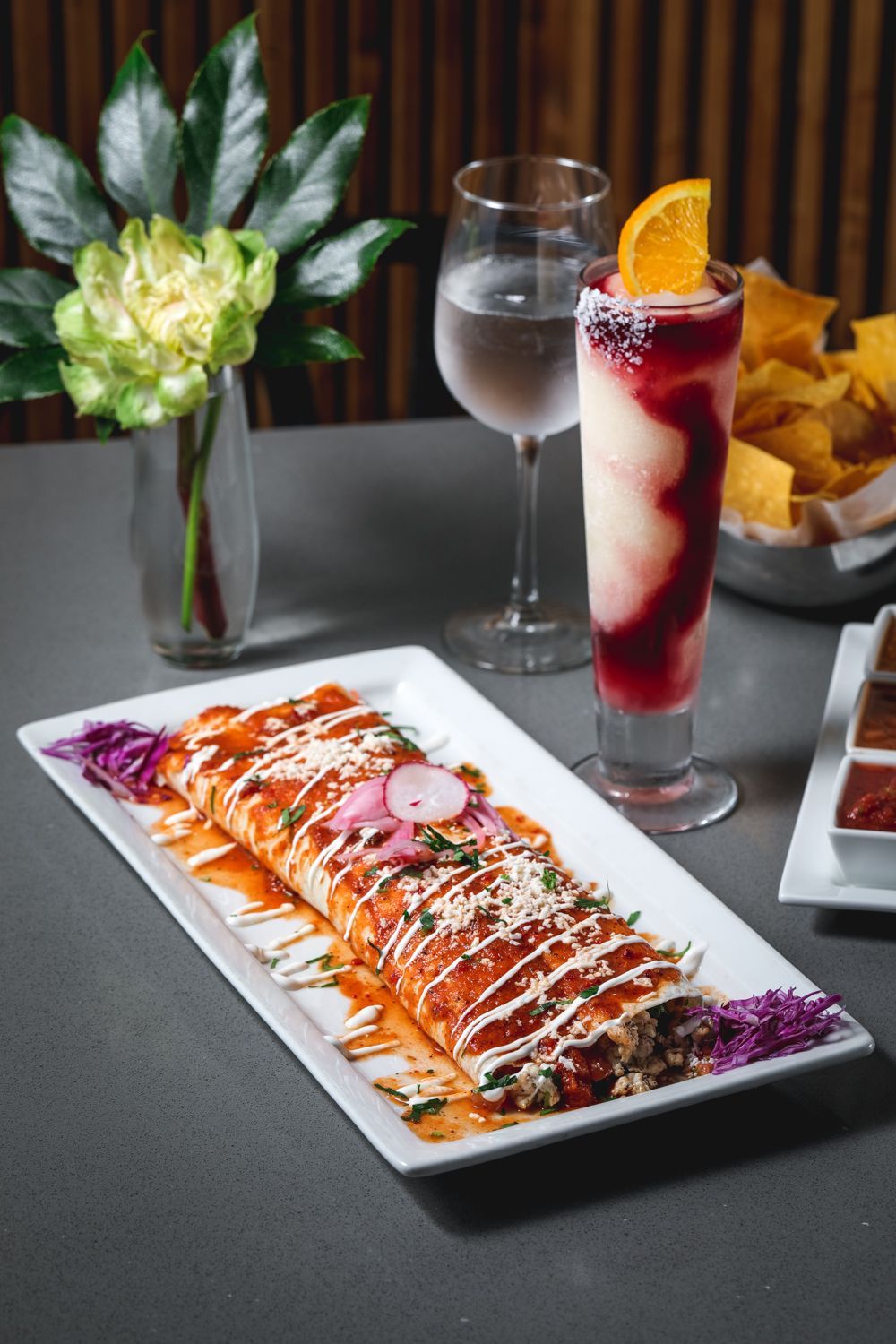 Burrito with sauce, drink with orange slice, water glass, chips, and plant on table.