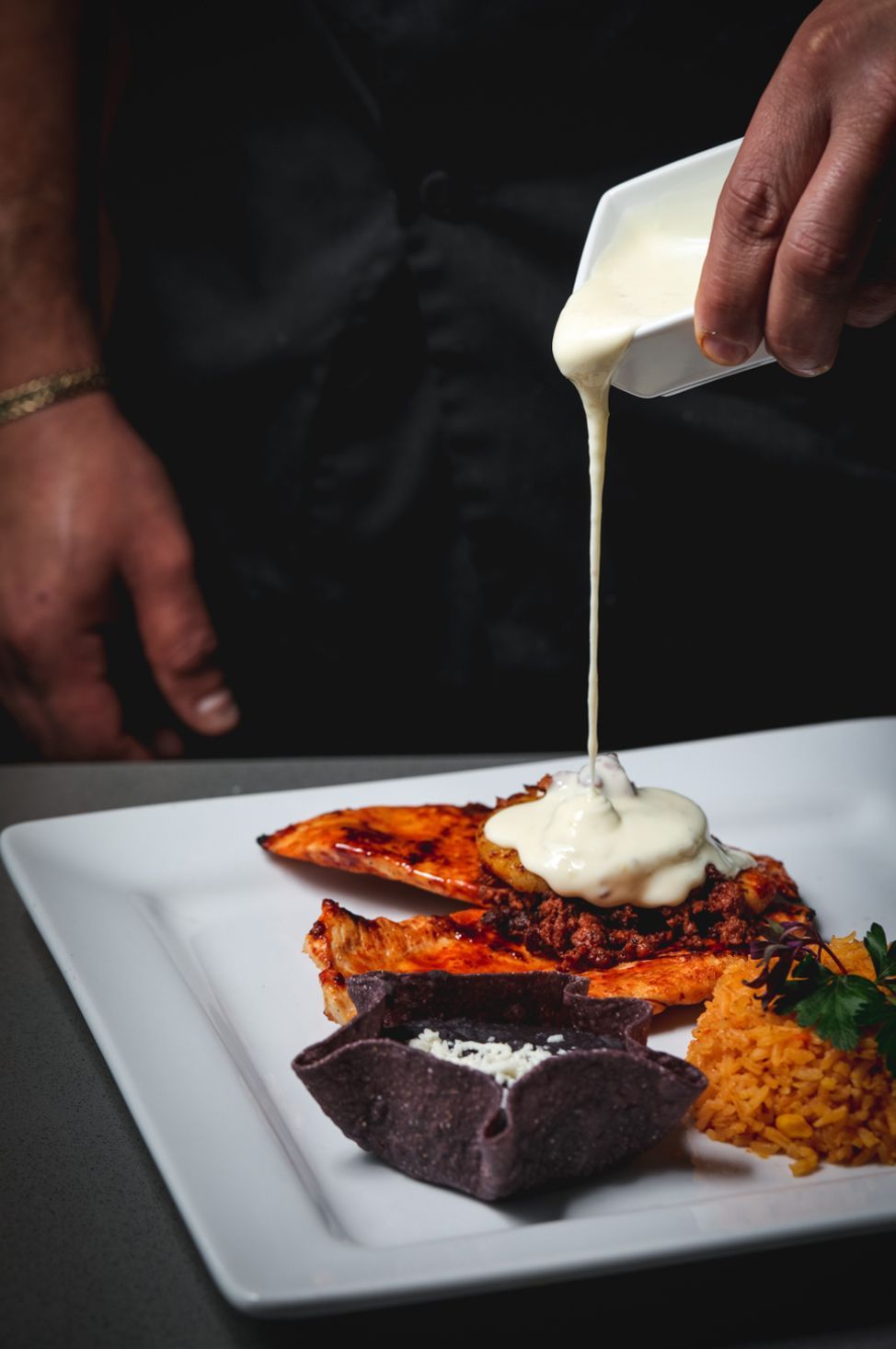 Chef pouring creamy sauce over a dish of food, plated on white, with a side of grains and a dark-colored edible bowl.