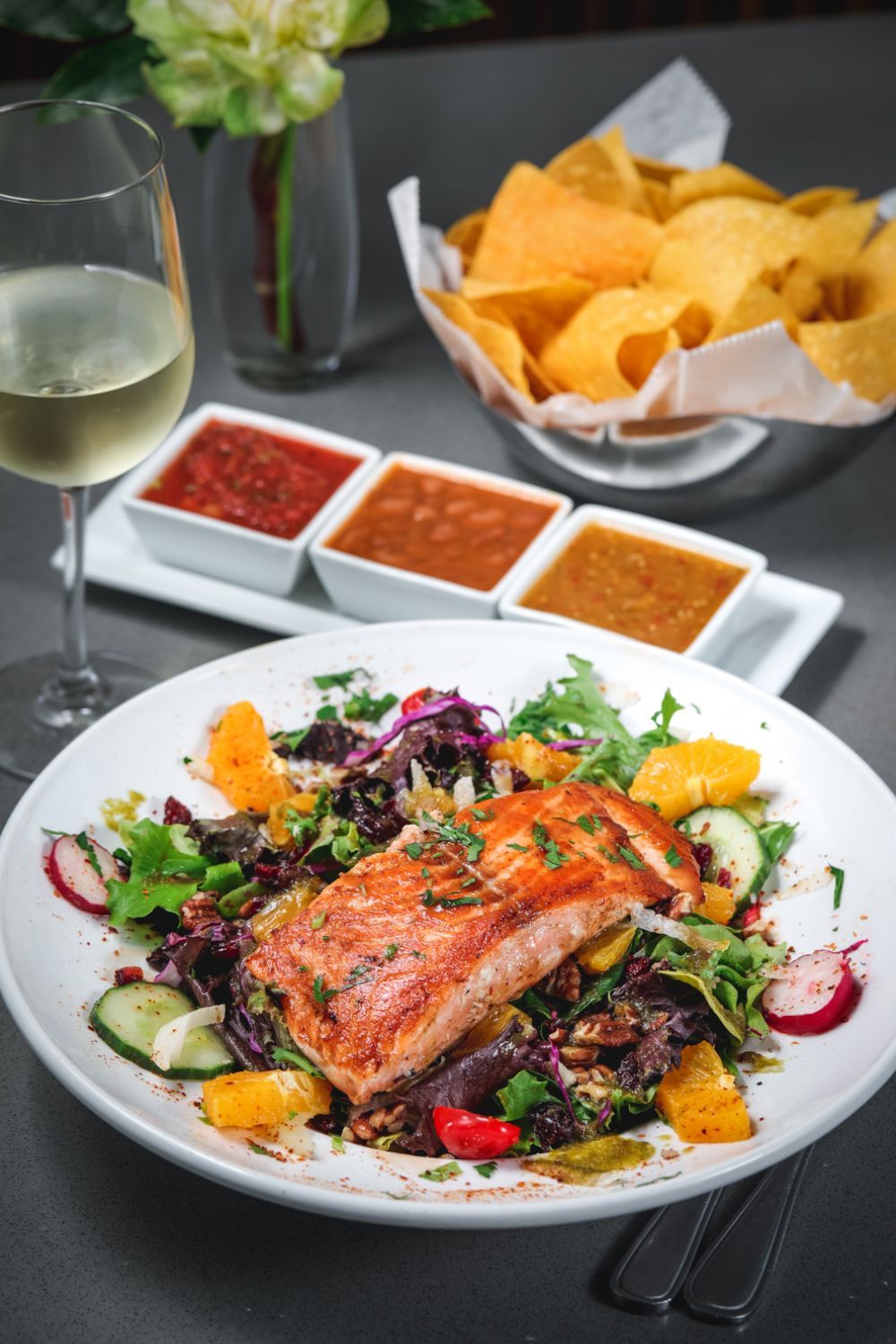 Grilled salmon salad, chips and salsa, wine glass on table.