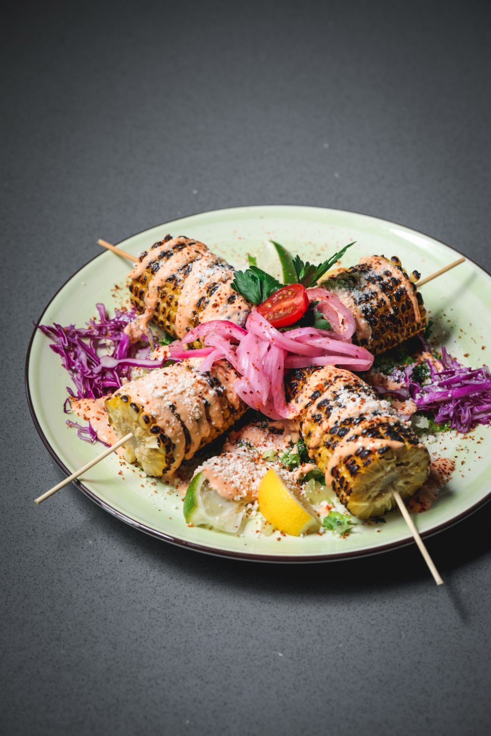 Grilled corn on the cob skewers with pink sauce, red cabbage, and pickled onions on a green plate.