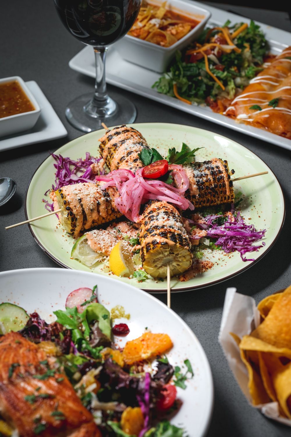 Grilled corn on a plate with other dishes: salad, enchilada, chips, sauces, and a glass of red wine.