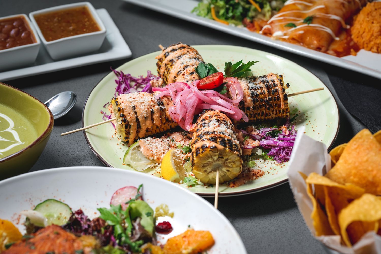Grilled corn on a plate with pickled onions, alongside other Mexican dishes like enchiladas and a salad.