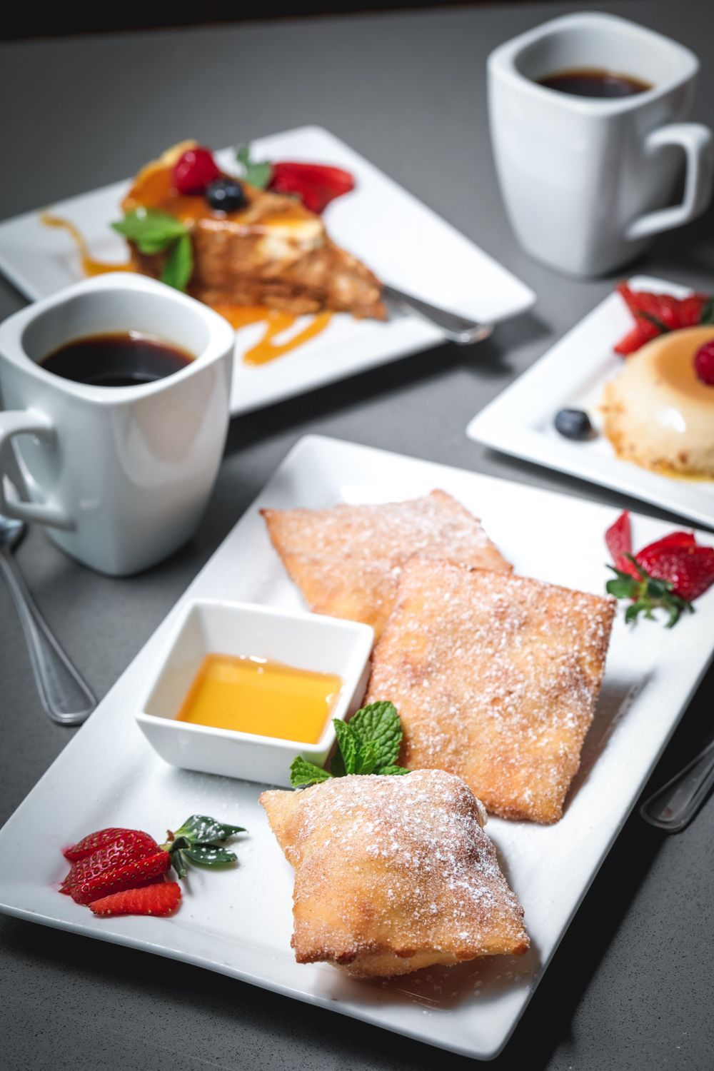 Plated dessert with powdered sugar, dipping sauce, and berries, alongside coffee cups.