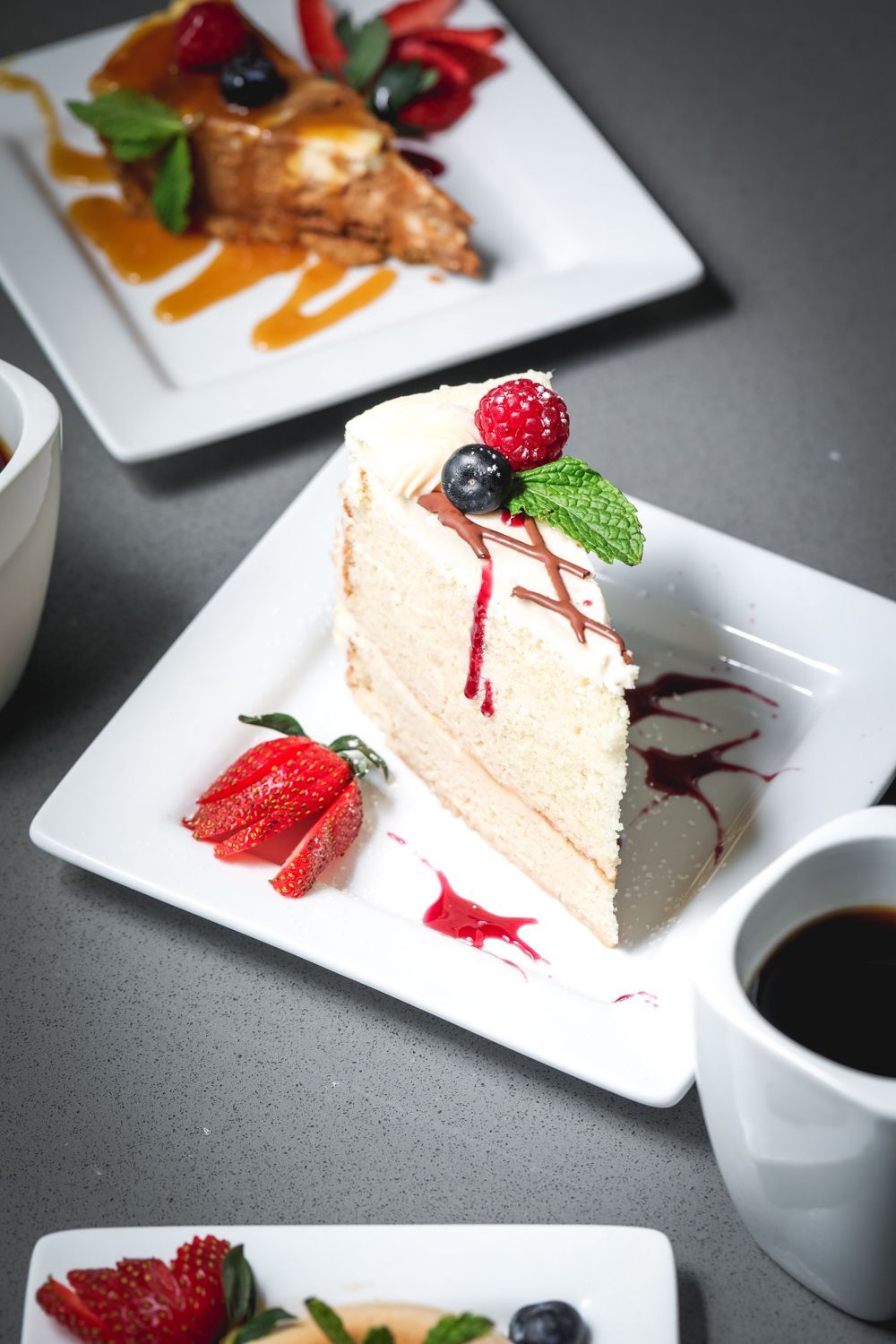 Slice of layered cake on a white plate with berries, syrup, and coffee.