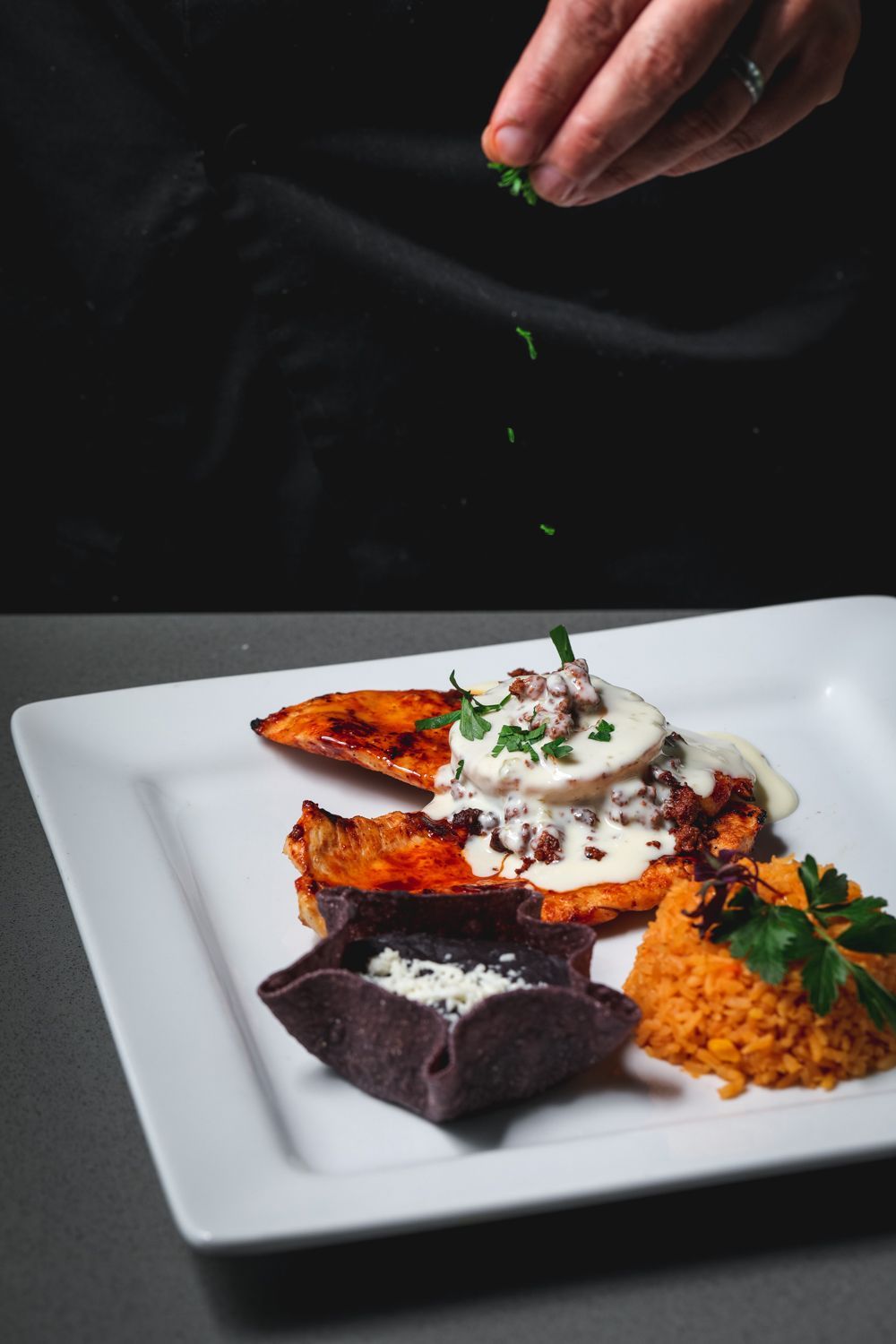 Chef garnishes plate with chicken, rice, and black tortilla basket filled with cream, on a white plate.