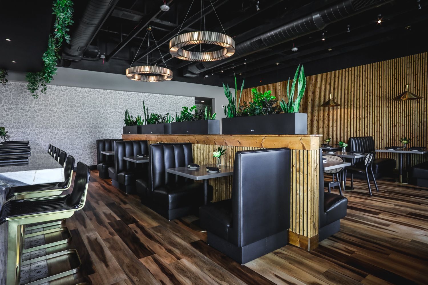 Restaurant interior with booths, bar seating, plants, and modern light fixtures.