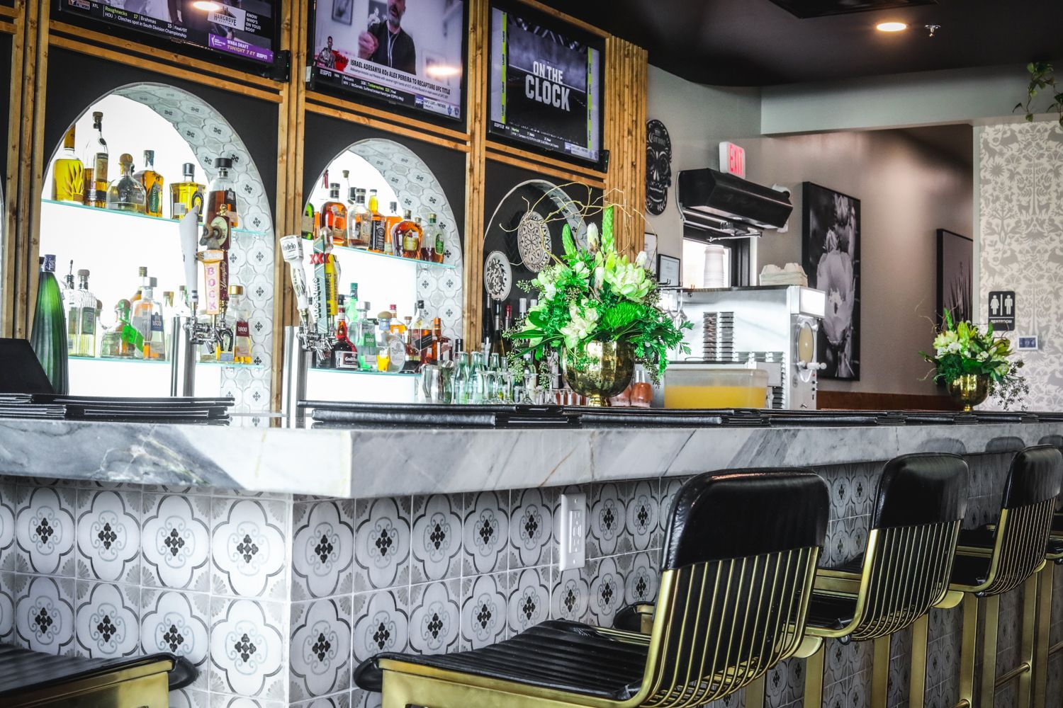 Bar with marble countertop, decorated front, and black and gold stools. Liquor bottles and TVs are visible.