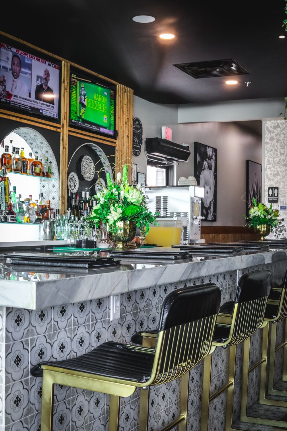 Bar interior with stools, marble countertop, decorative tiled wall, TVs, and liquor bottles.