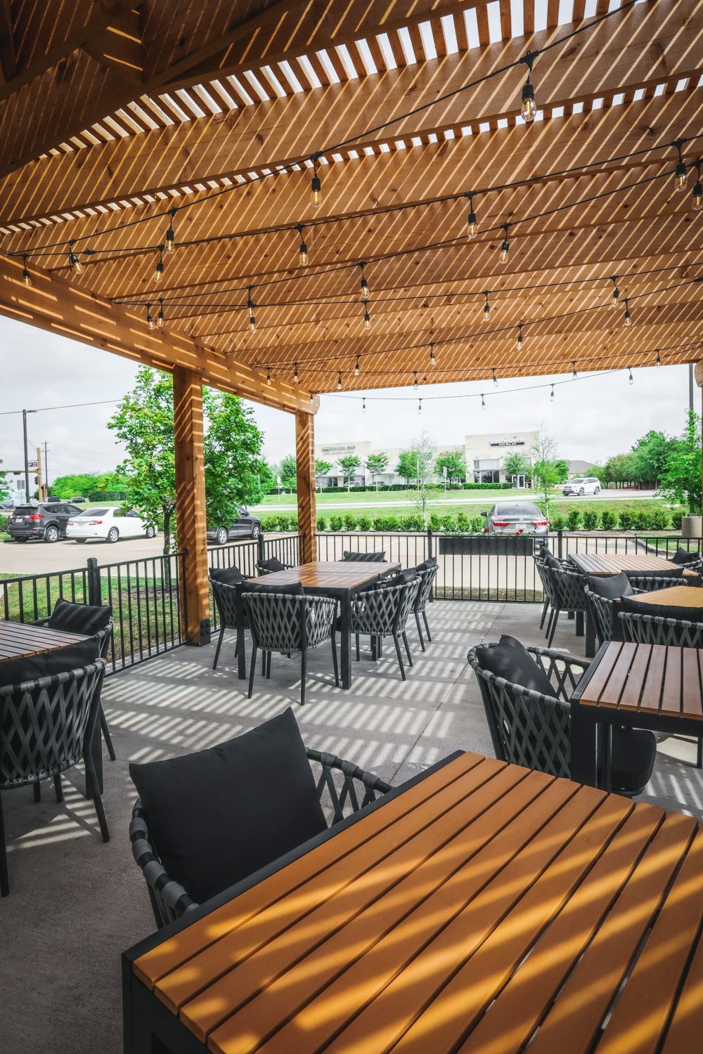 Outdoor patio with tables and chairs under a wooden trellis; string lights, cityscape in the background.