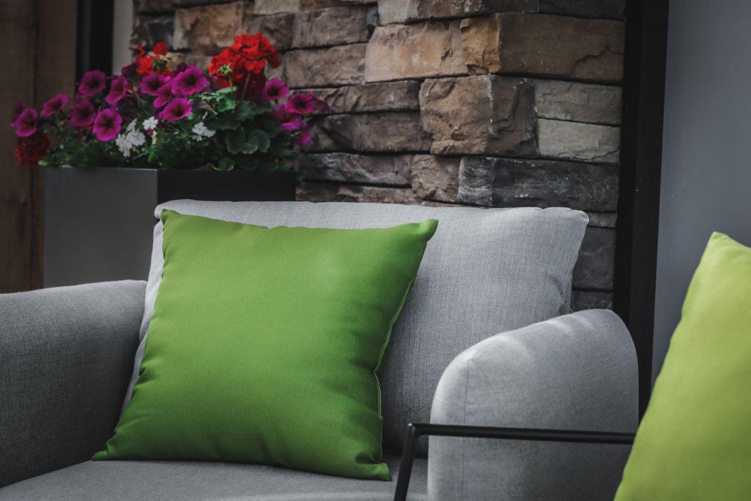 Green pillows on a gray outdoor chair, with red and purple flowers in a planter, stone wall background.