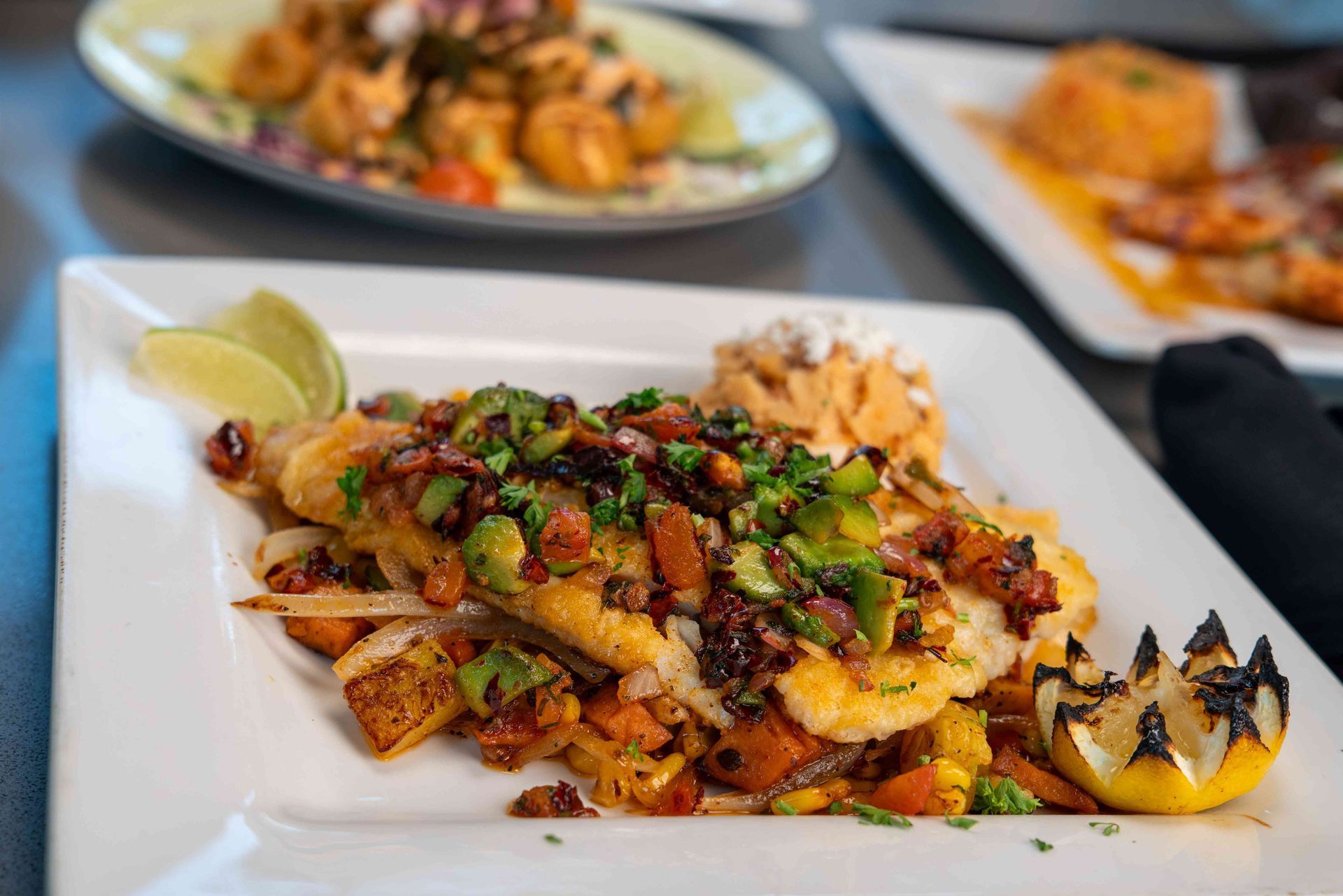 Plate of pan-seared fish topped with avocado salsa, served over roasted vegetables, with lime wedges.