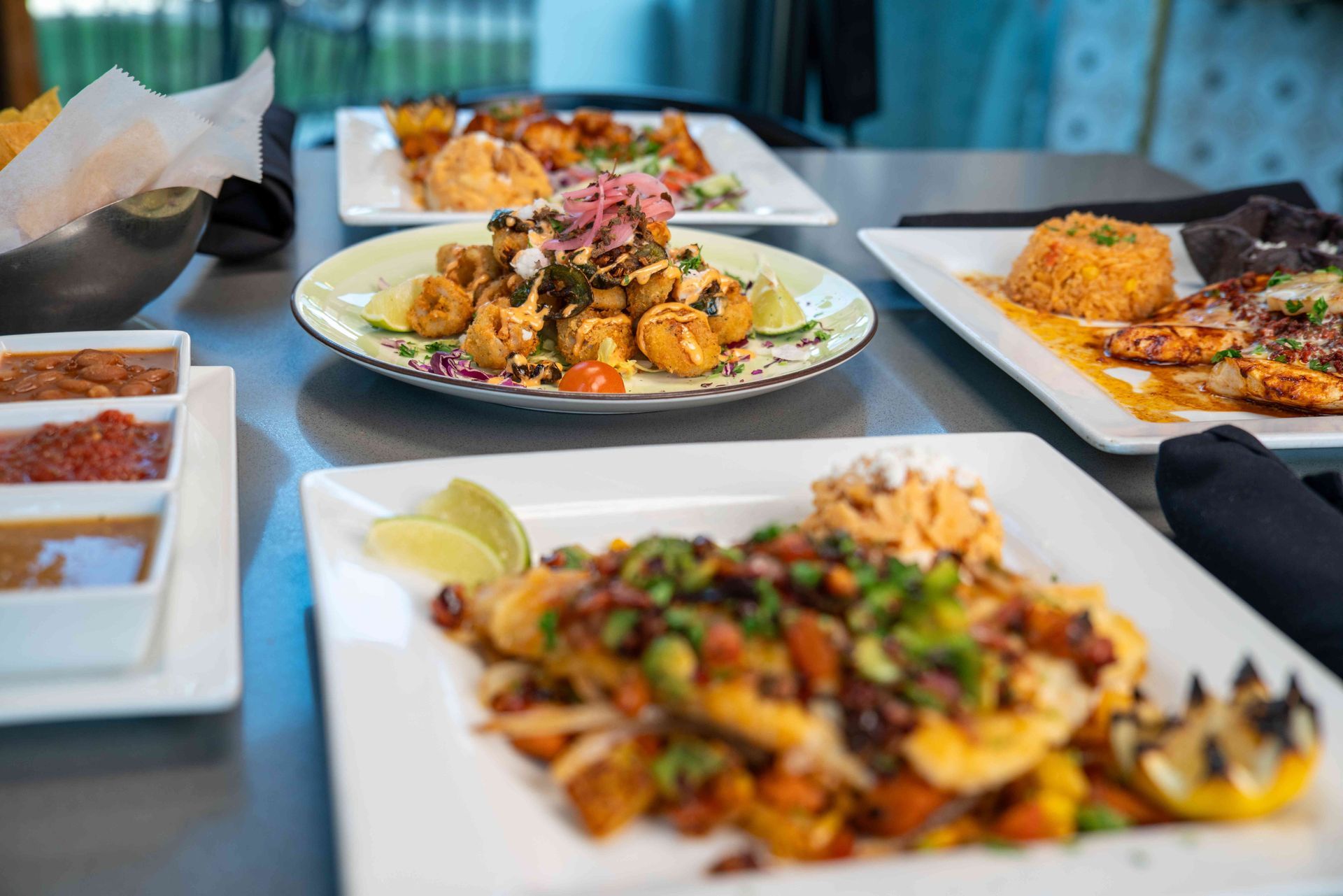 Various Mexican dishes on a gray table: fried foods, rice, fish, salsa, and limes.
