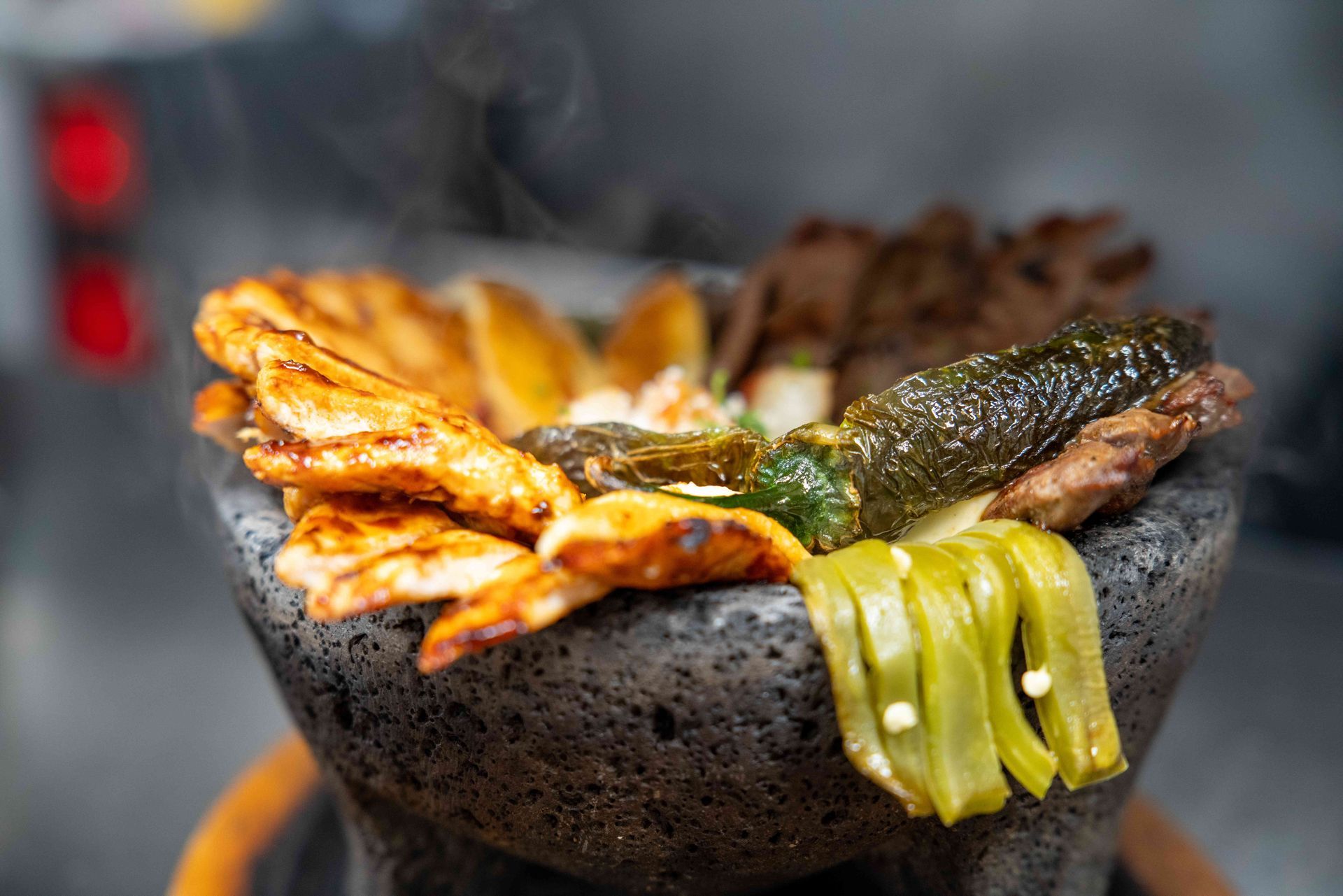 Hot molcajete bowl filled with grilled meats, vegetables, and peppers; steam rising.