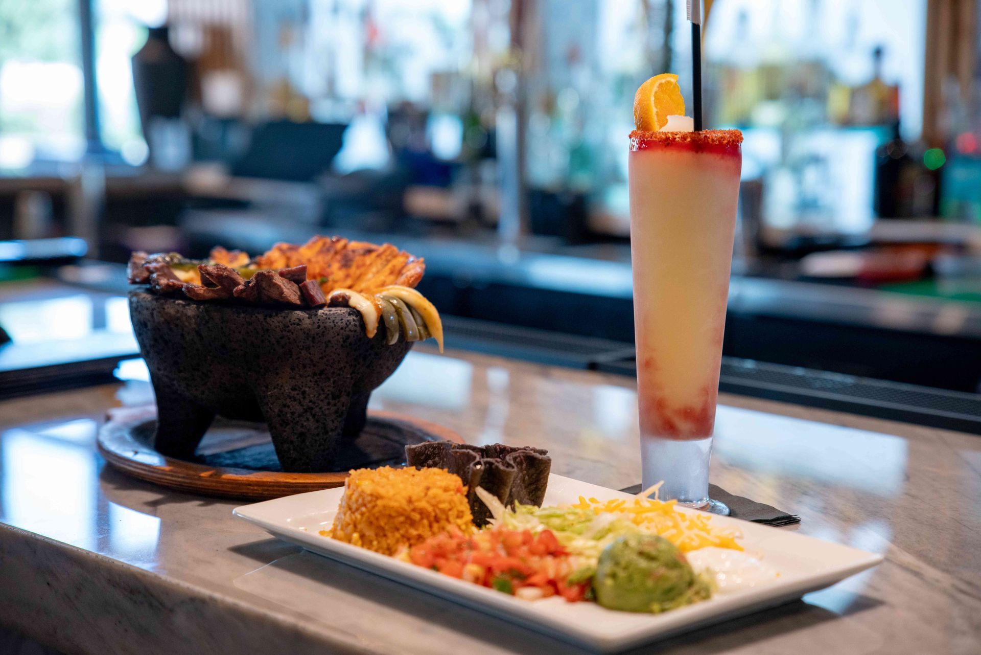 Mexican dish with grilled meat in a molcajete, side of rice, beans, and cocktail on a bar.