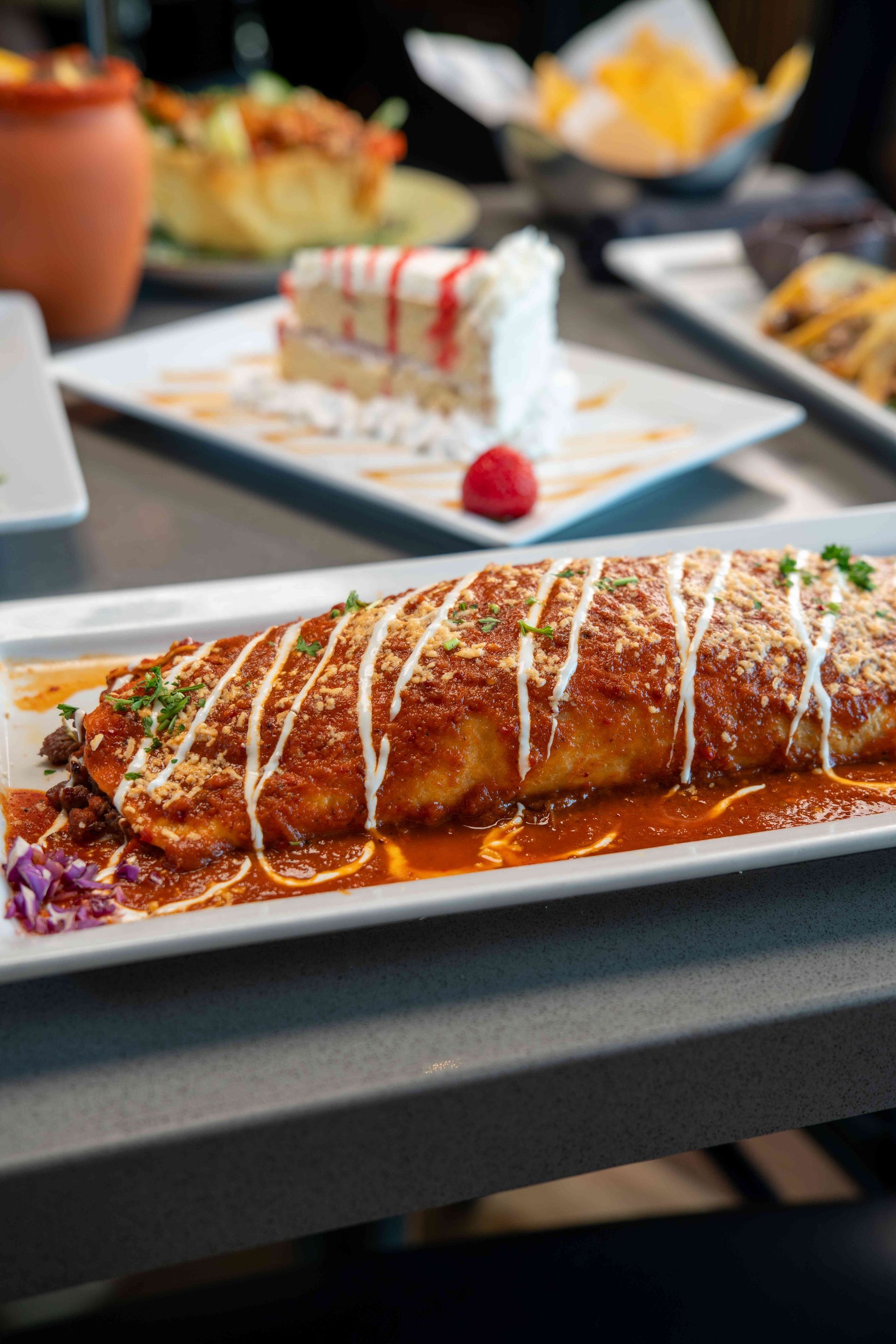 Burrito covered in sauce, drizzled with cream, on a rectangular plate with other Mexican dishes in the background.