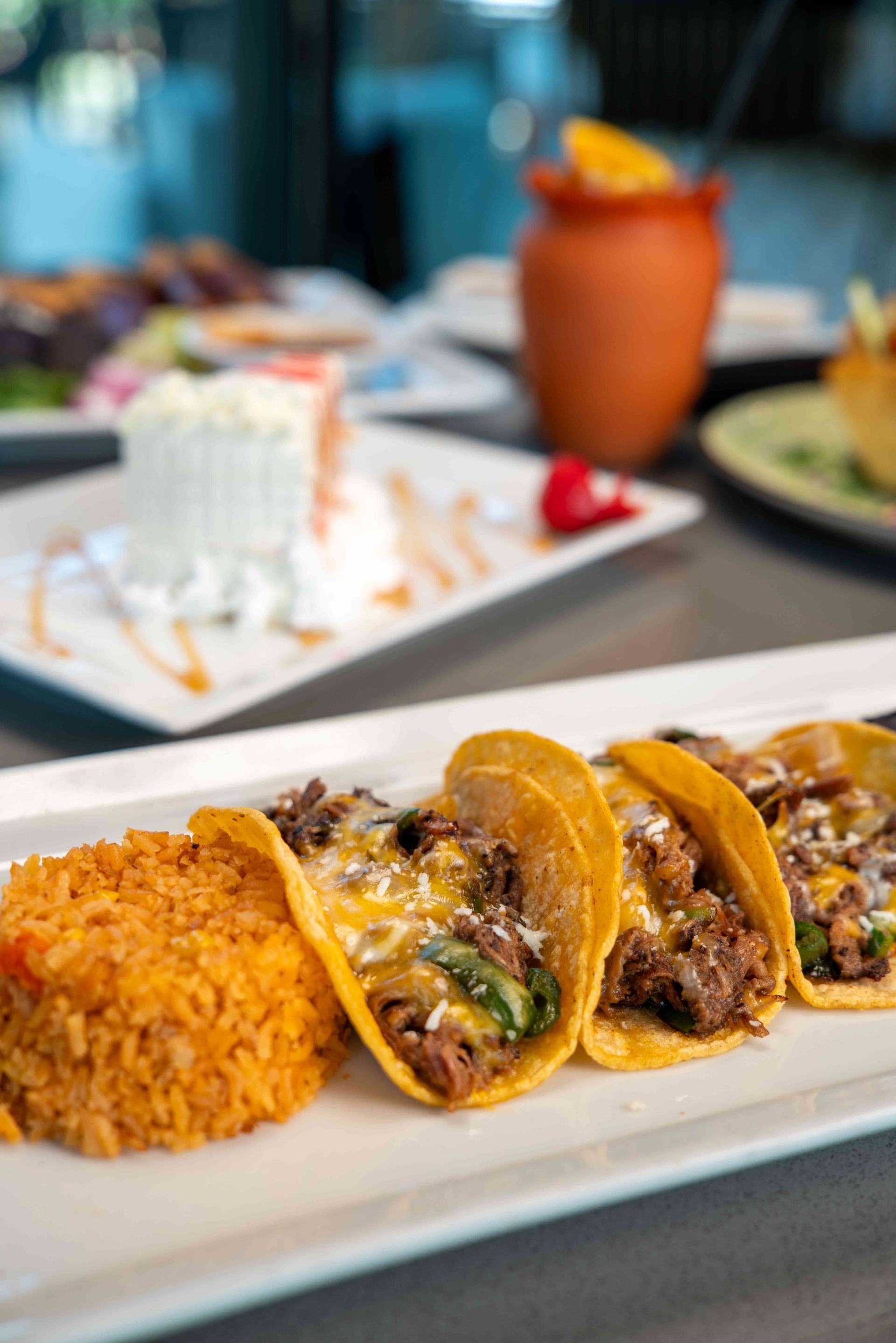Tacos with rice on a white plate, dessert with caramel drizzle, and a clay mug with orange garnish in the background.