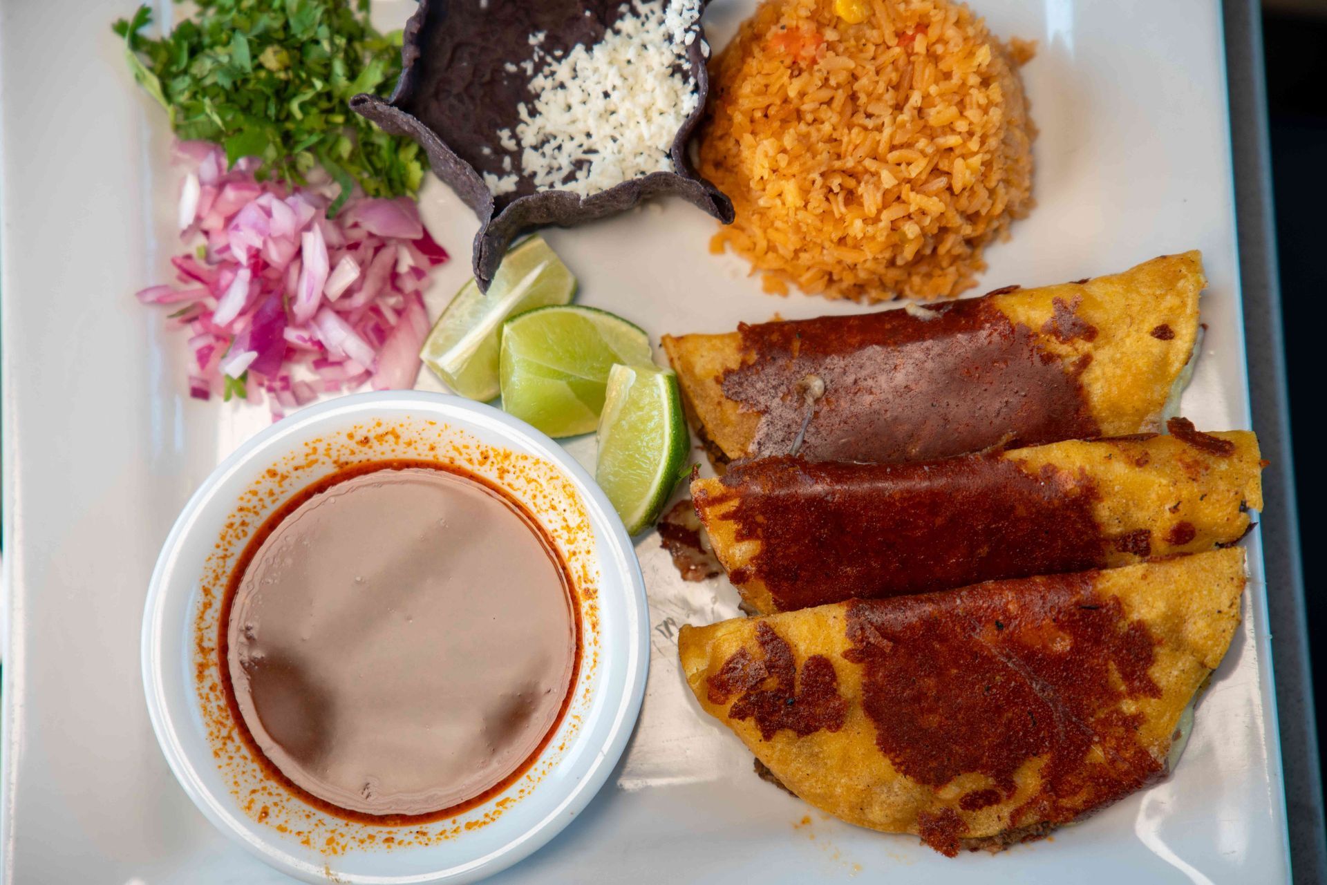 Plate of tacos, rice, and toppings: cilantro, onions, sauce, blue tortilla, and limes.