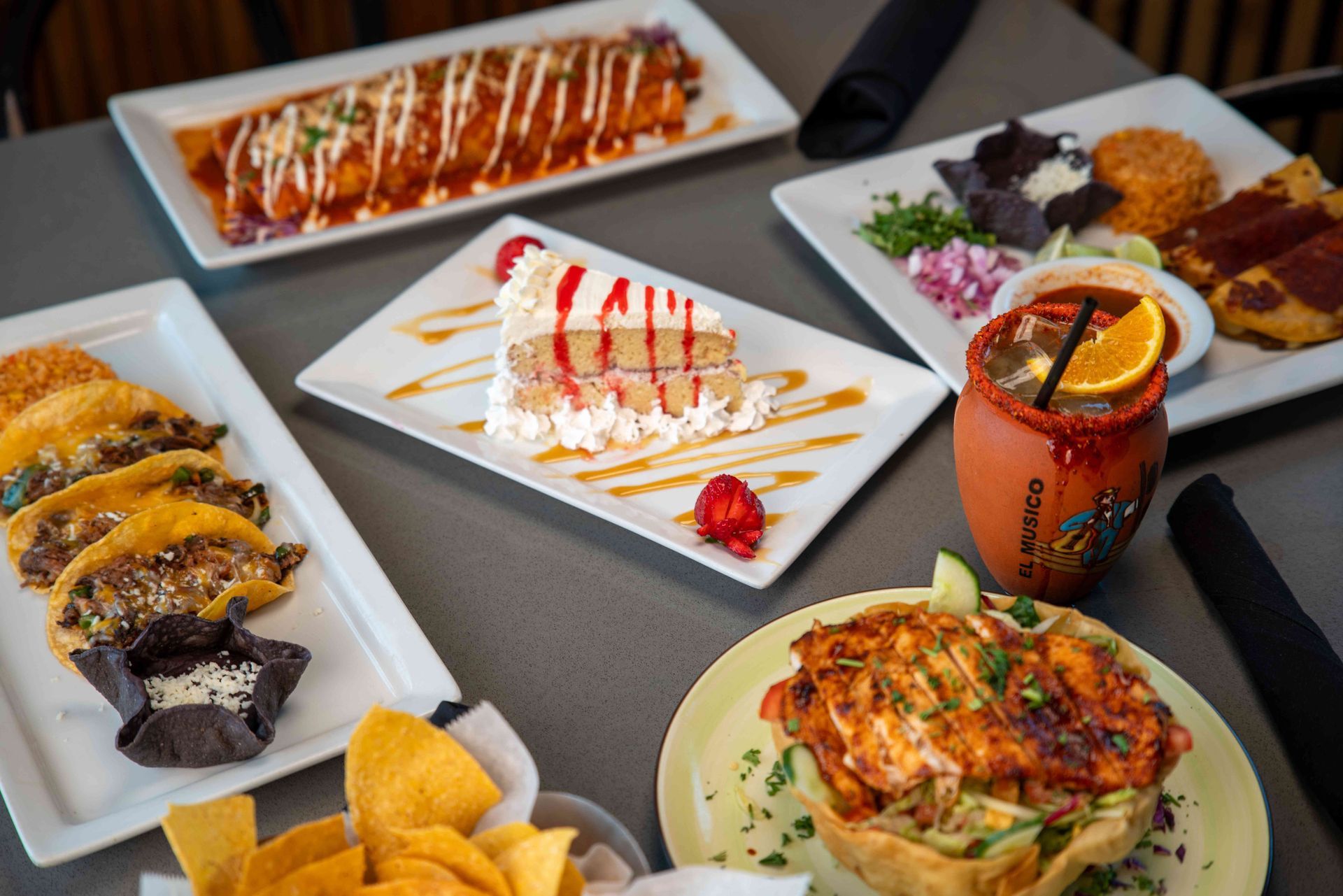 Mexican food spread on a table: enchiladas, tacos, cake, salad, drink, and chips.