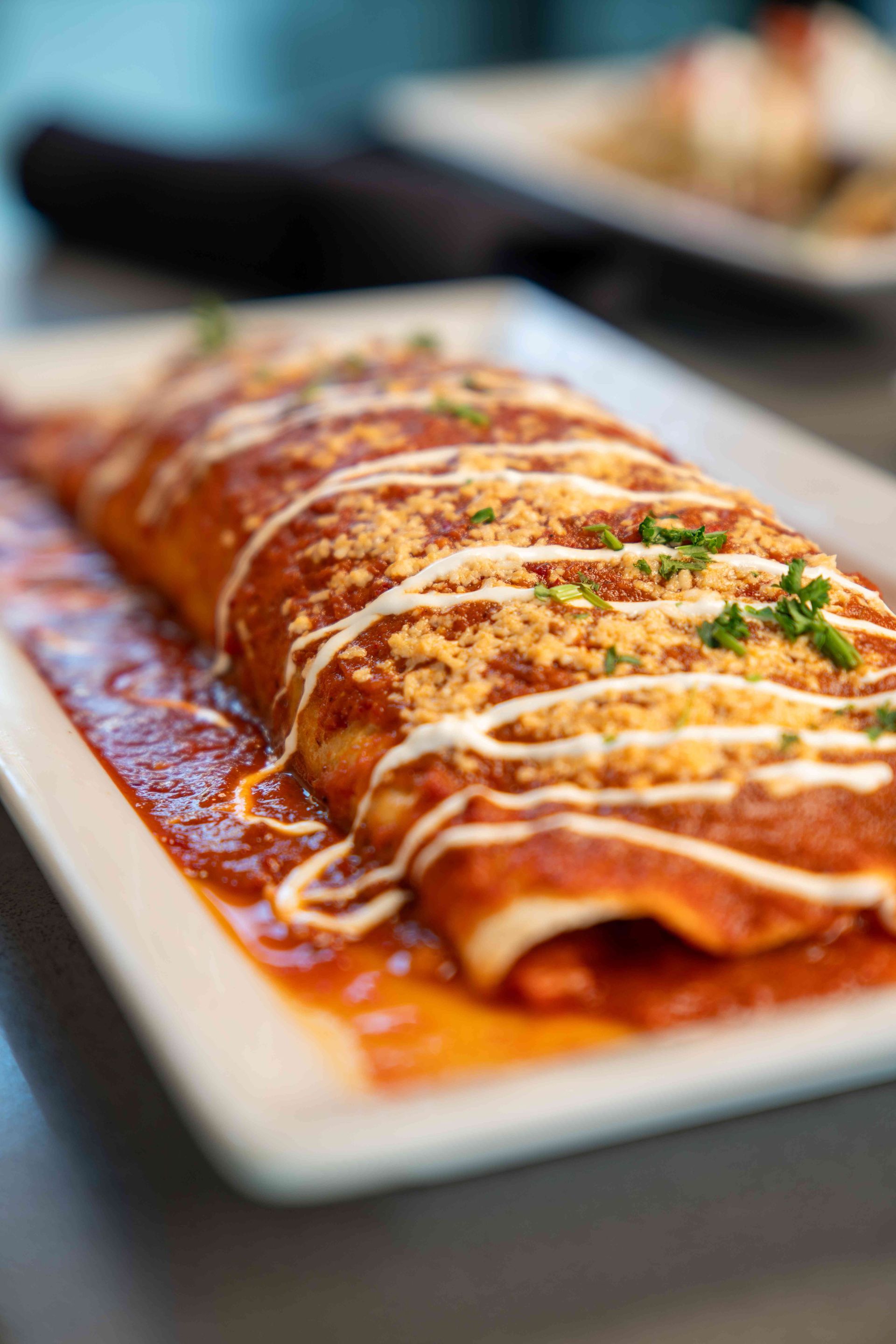 Enchilada covered in red sauce and drizzle, on a white plate. Garnished with cheese and chives.