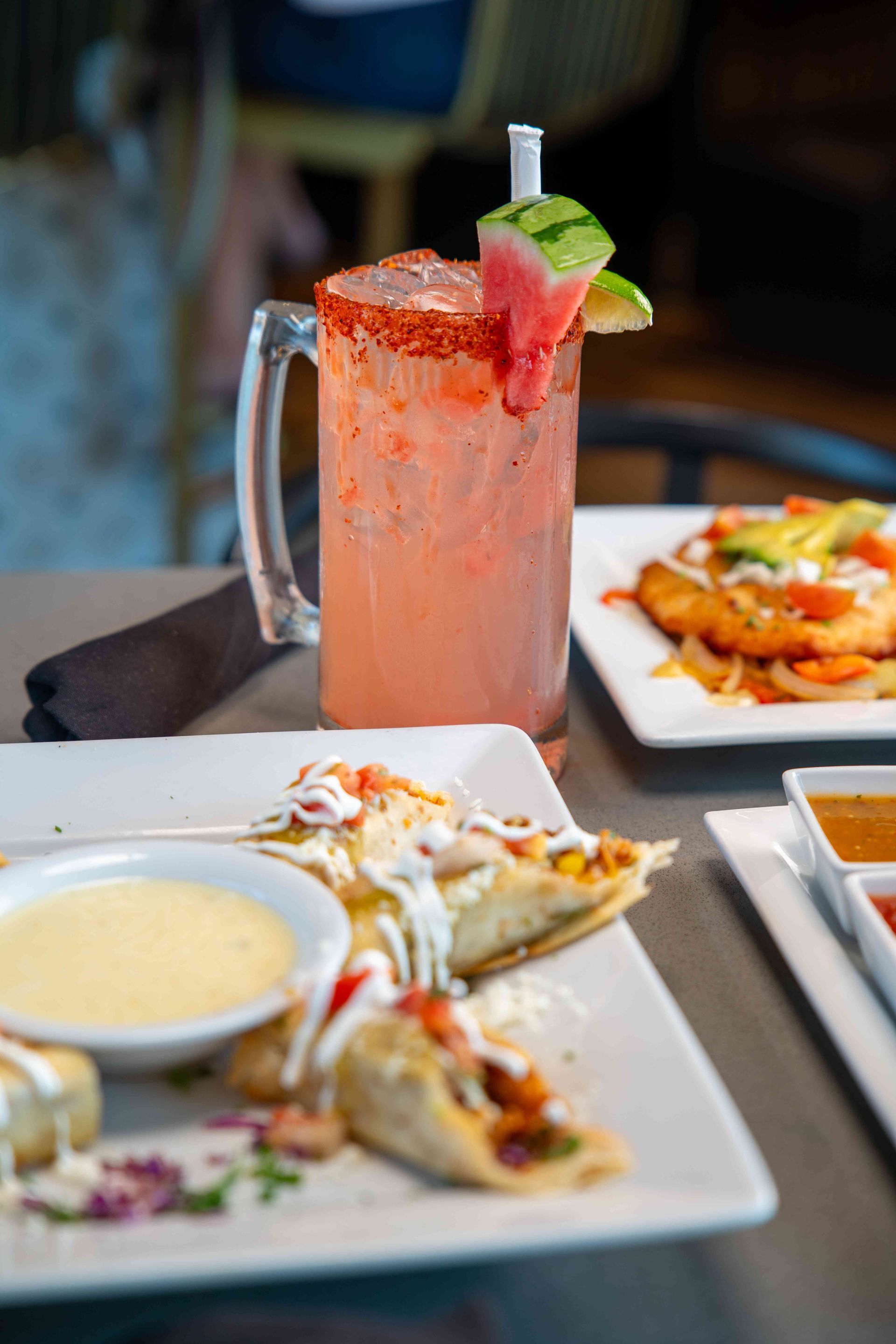 Pink cocktail with watermelon garnish next to appetizer plates on a table.
