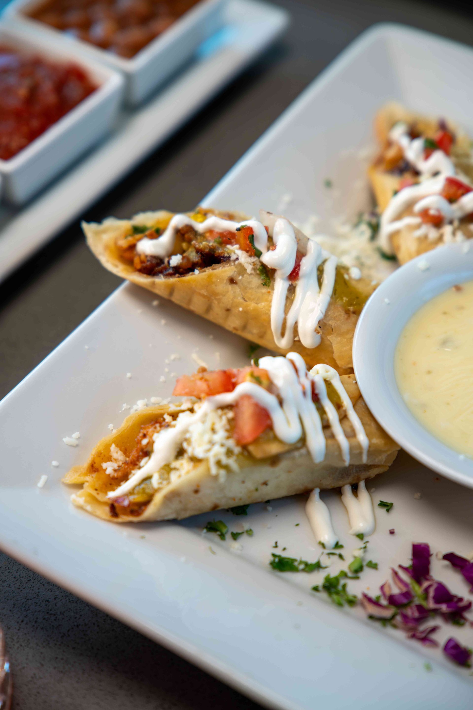 Tacos on a white plate, drizzled with white sauce and topped with tomatoes and cheese, with a side of yellow sauce.