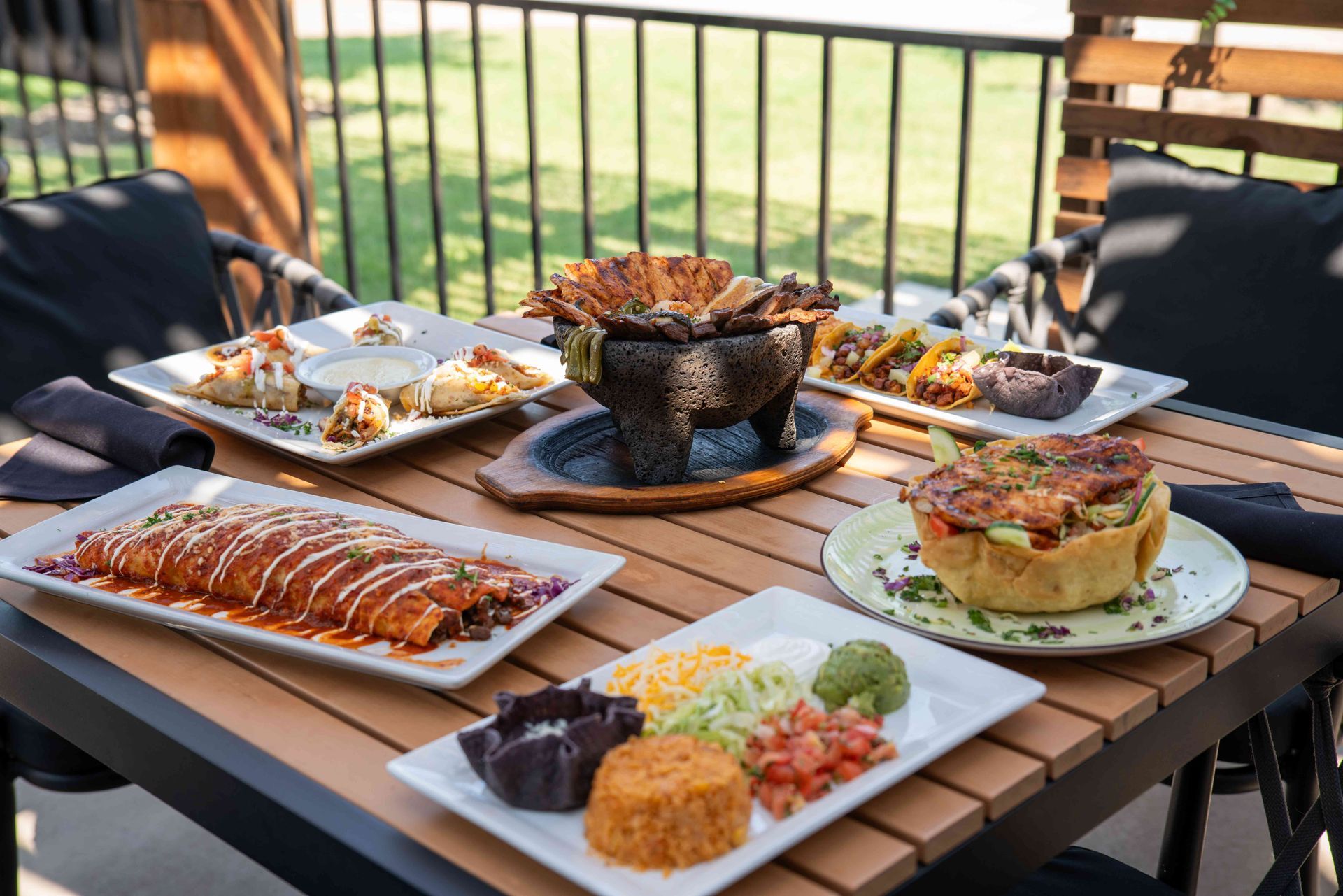 Assorted Mexican dishes on a wooden outdoor table: enchiladas, tacos, rice, and guacamole.