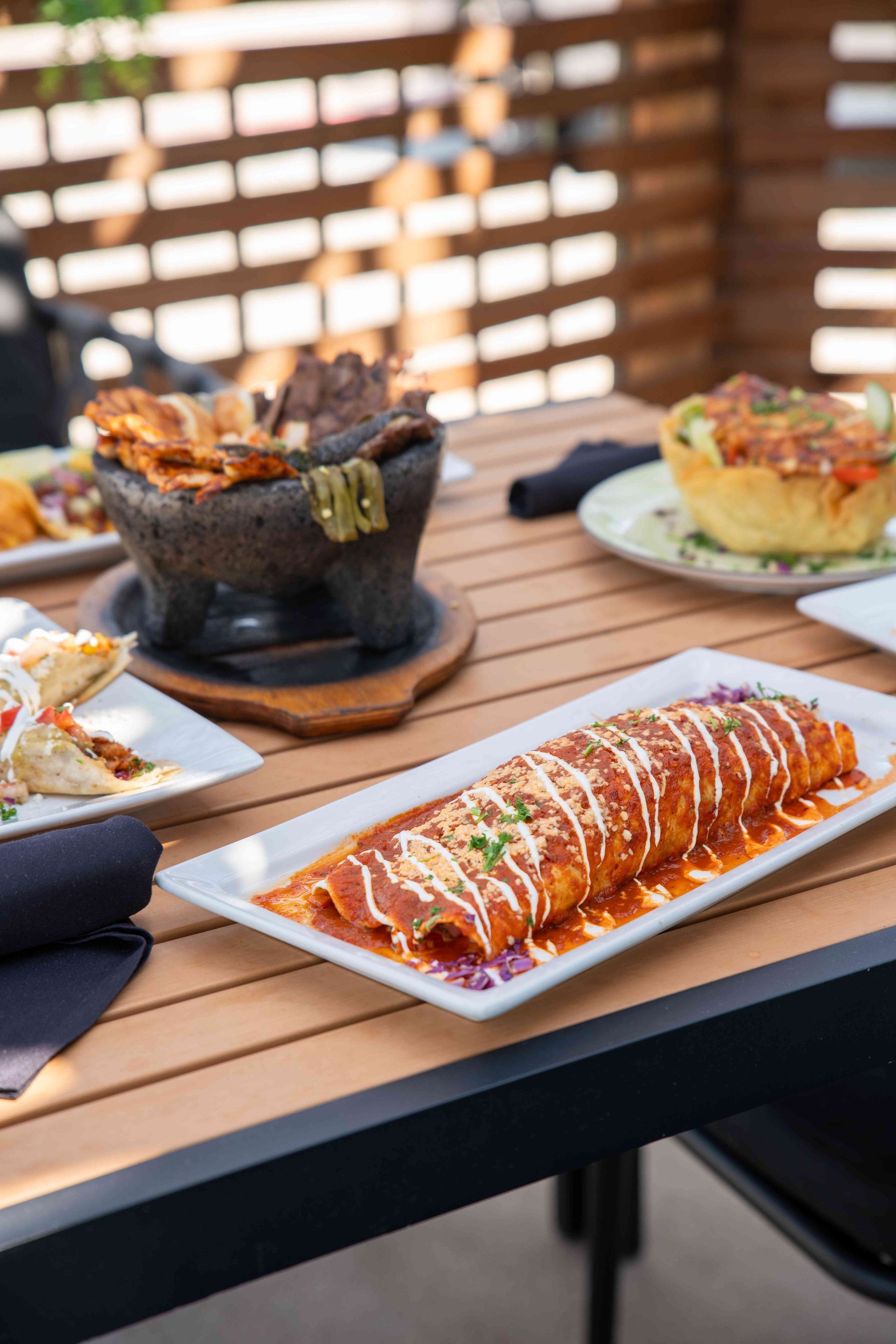 Mexican food dishes on a wooden table outdoors. Enchilada, molcajete, and other plates visible.