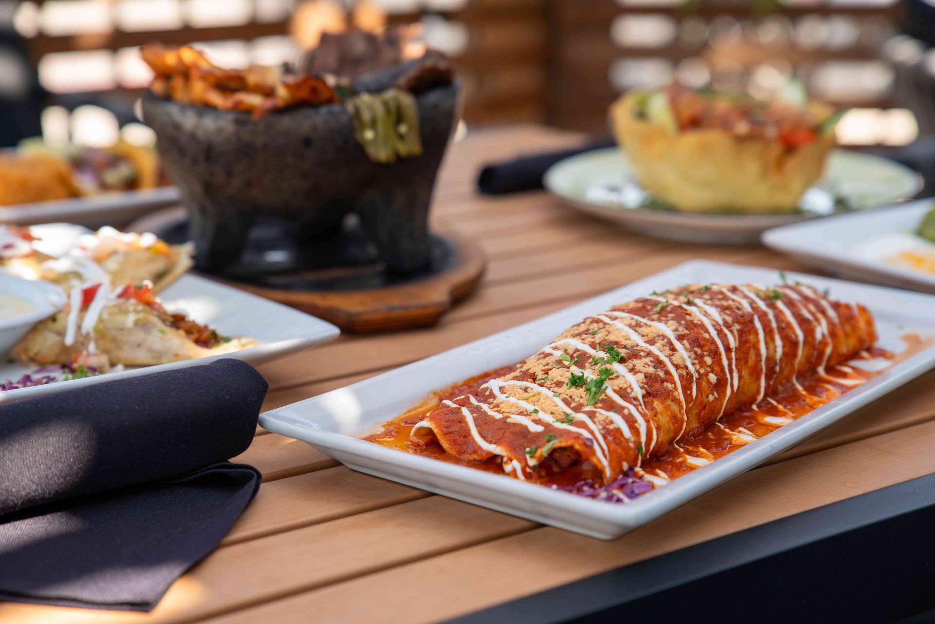 Burrito covered in red sauce and drizzle, on a rectangular plate, with other Mexican dishes on a wooden table.