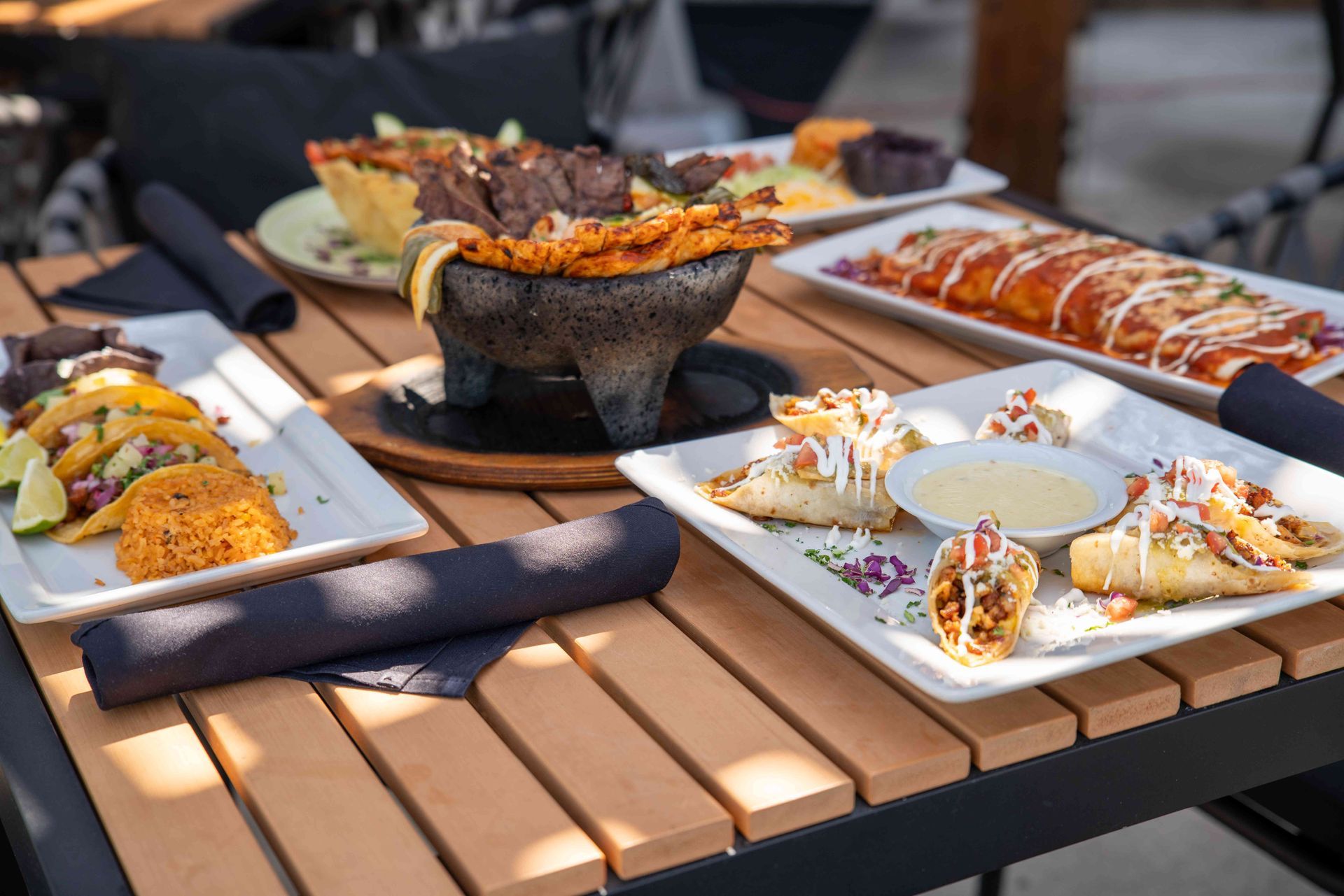 Mexican food dishes on a wooden table outdoors. Tacos, enchiladas, and a molcajete are visible.