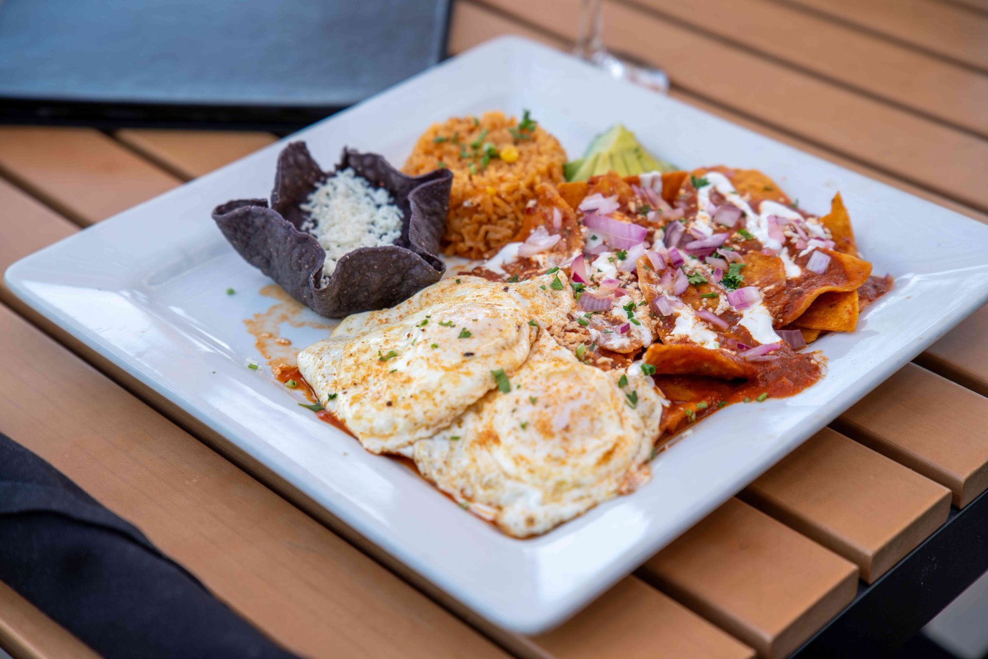 Plate of Mexican food: eggs, chilaquiles, rice, black tortilla bowl, and garnish on a wooden table.