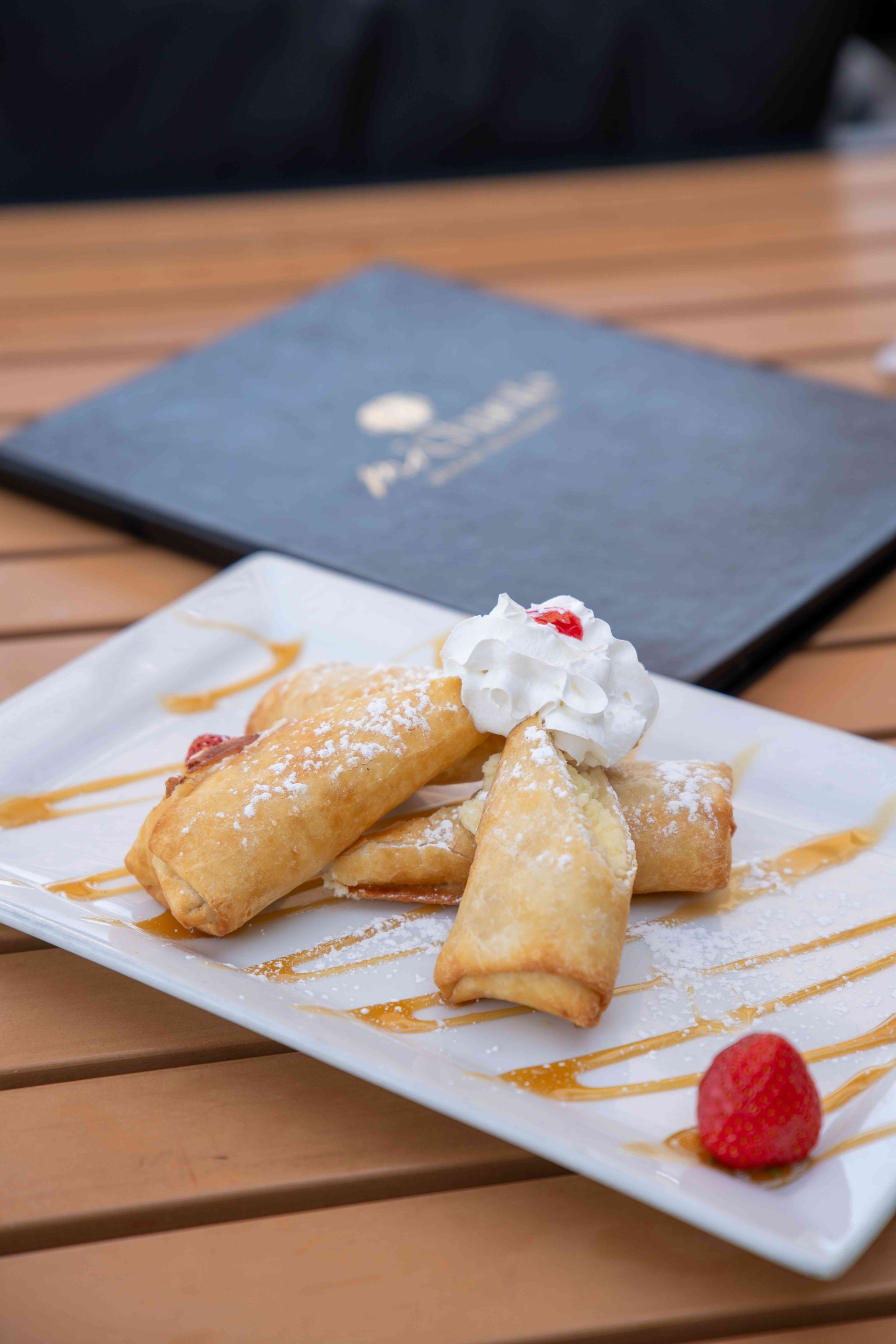 Fried dessert rolls on a white plate with whipped cream, syrup, and a strawberry. Menu in the background.