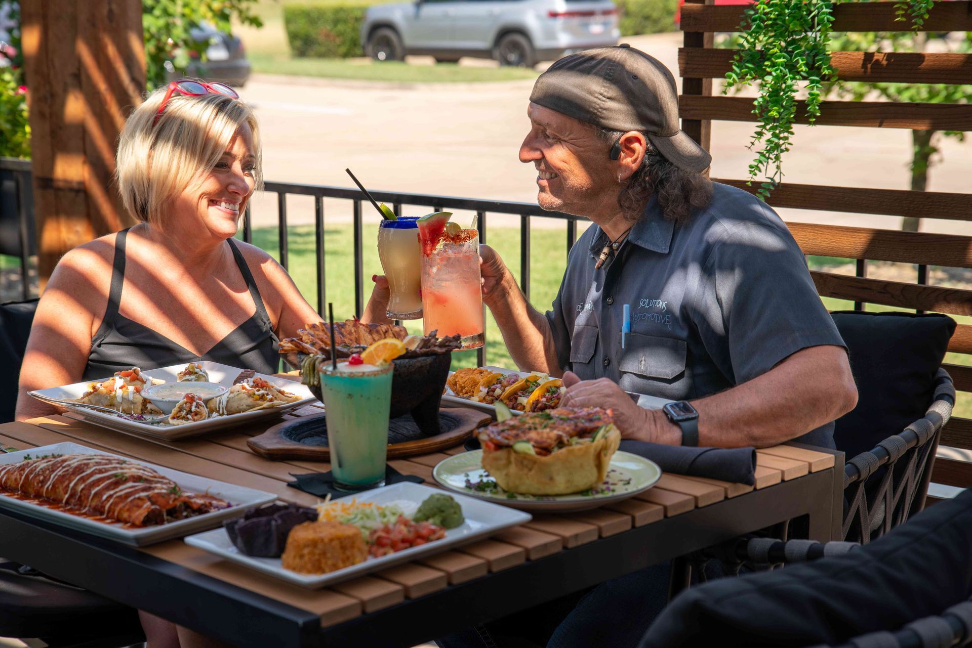 Couple toasting drinks at an outdoor restaurant table with food, sunny day.