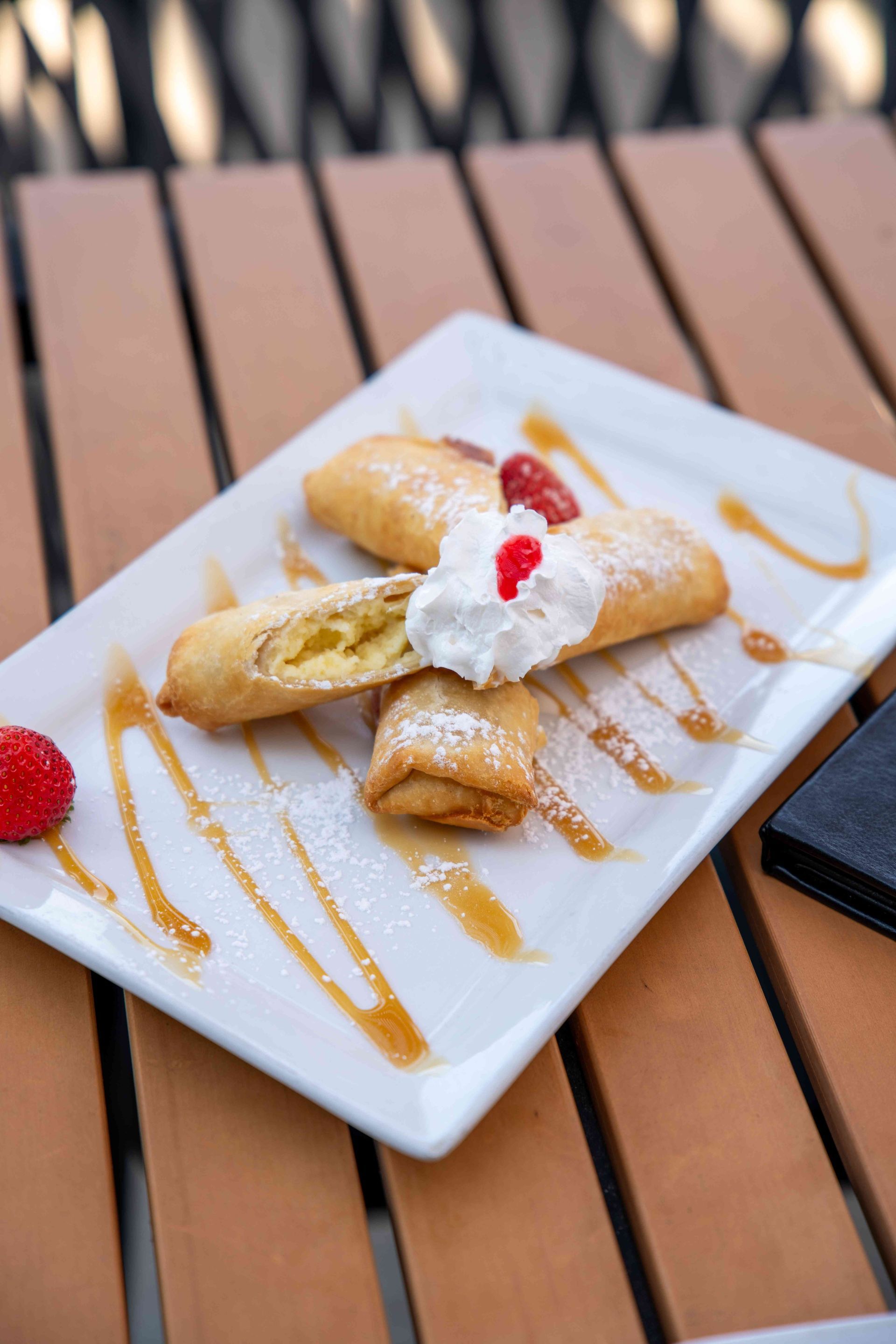 Fried pastry rolls with whipped cream, caramel drizzle, and raspberries on a white plate on a wooden table.