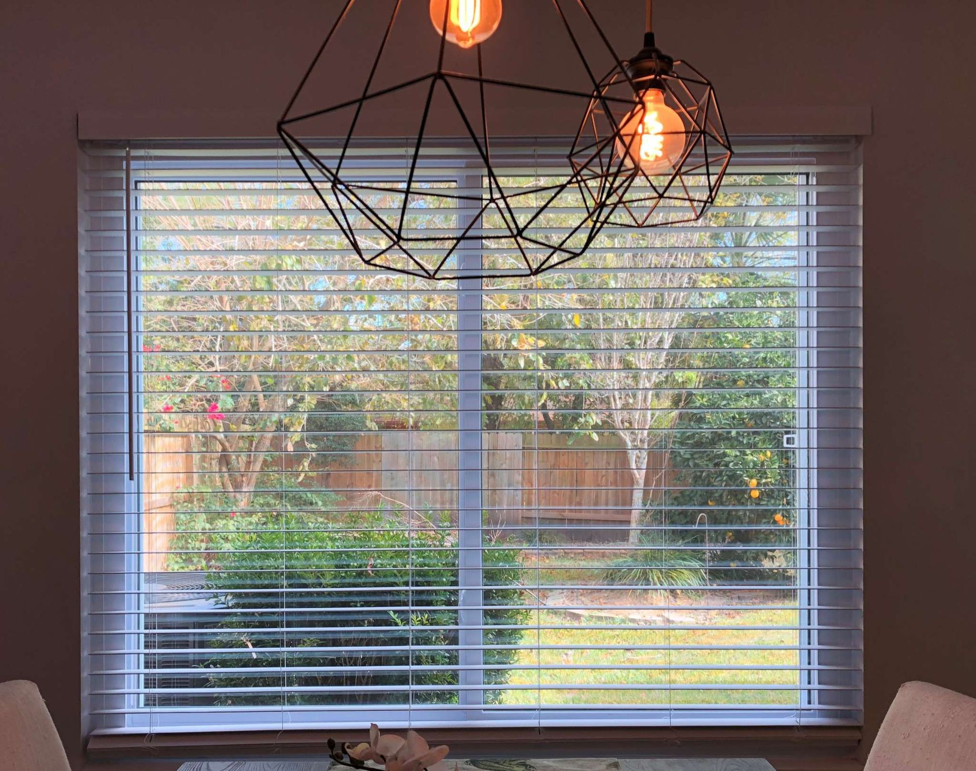 A dining room with a large window with blinds and a chandelier hanging from the ceiling.