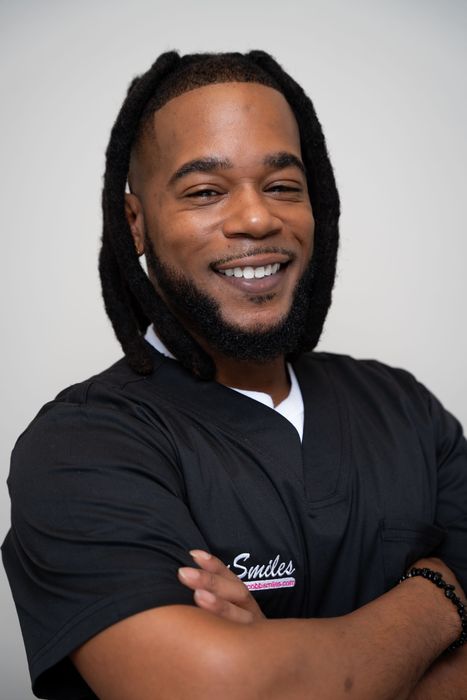 A man with dreadlocks and a beard is wearing a black scrub top and smiling.