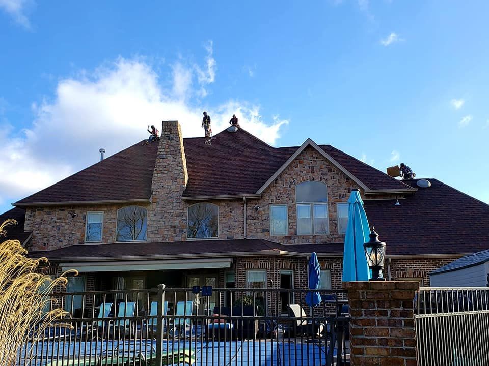 Workers are on the roof of a brick house on a sunny day. The house has a pool and blue umbrella in the backyard.