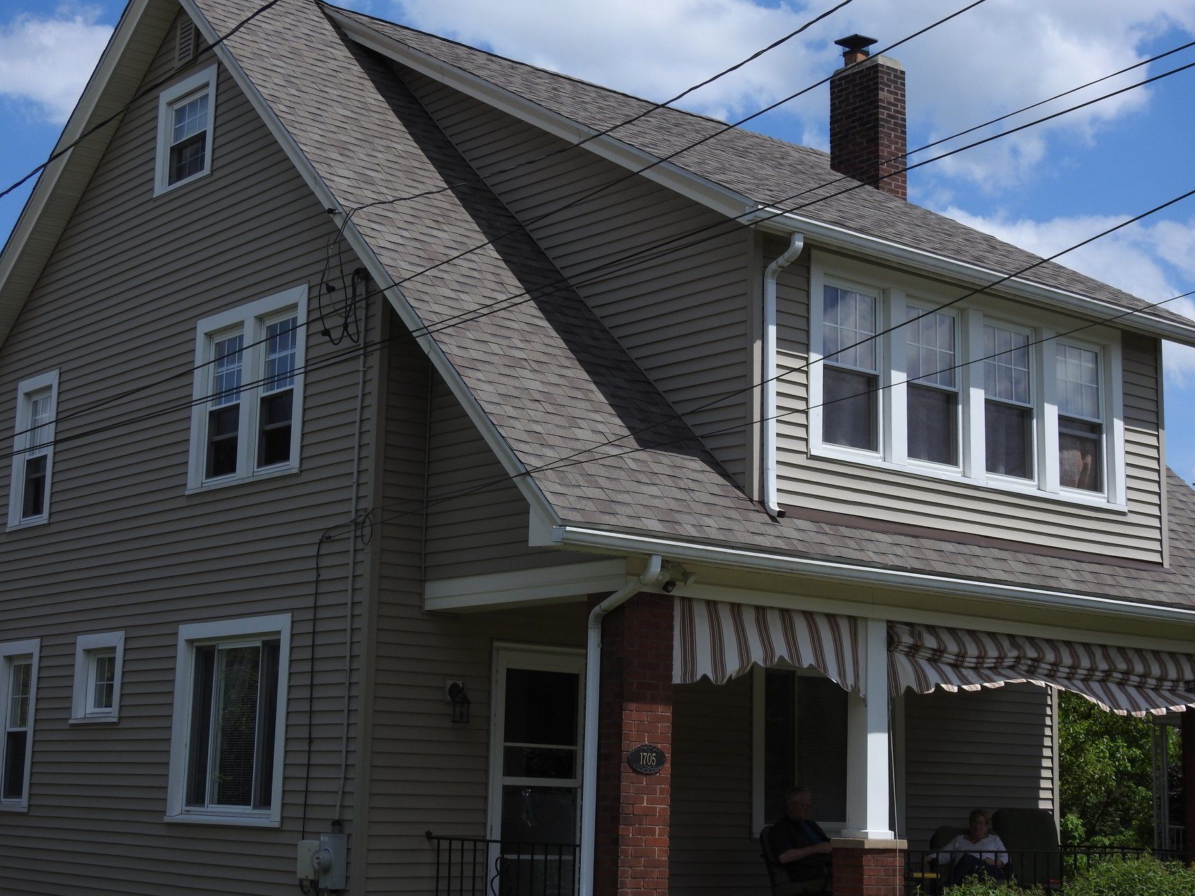 Two-story house with tan siding and a porch. Gray roof, white-framed windows, and striped awning.