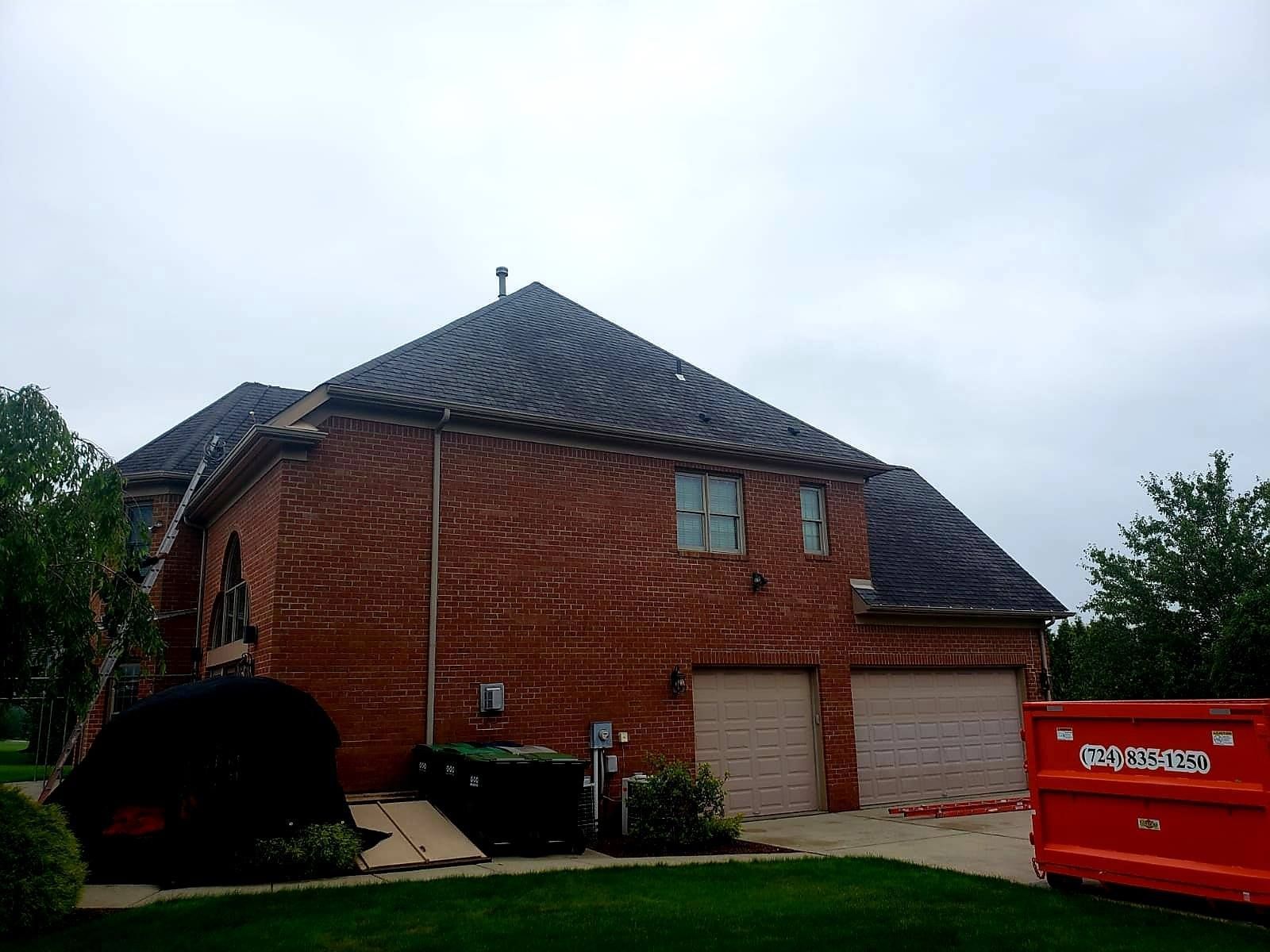 Red brick house with a dark roof and two-car garage under a cloudy sky. Construction debris and a dumpster are visible.