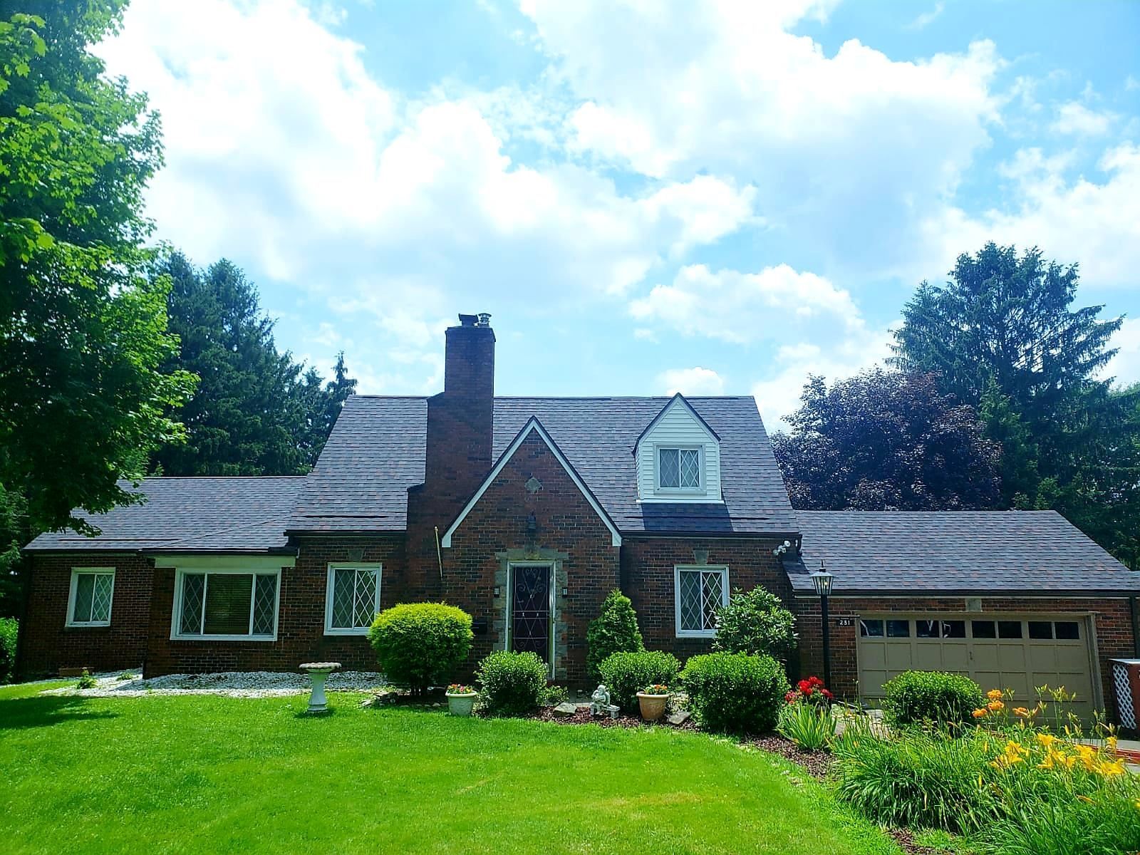 A brick house with a dark roof, chimney, and attached garage. Green lawn with bushes and trees against a bright blue sky with clouds.