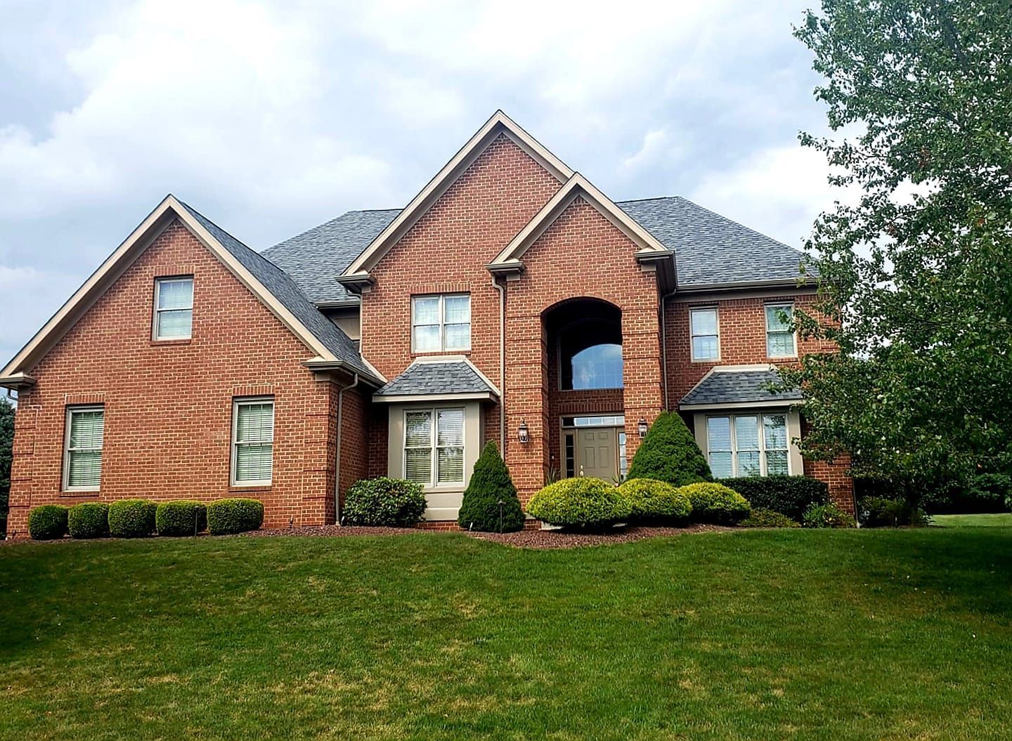 A two-story brick house with a dark shingled roof, set on a green lawn under a cloudy sky.