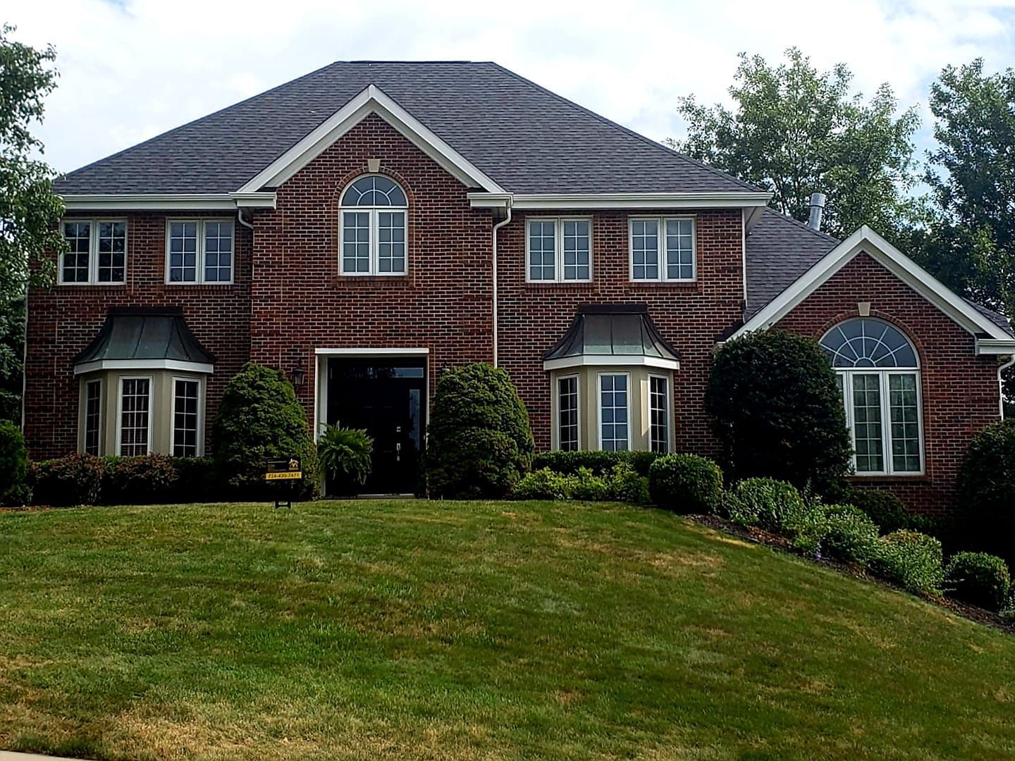 Two-story brick house with a dark gray roof and several windows. Green lawn and shrubbery in the front yard.