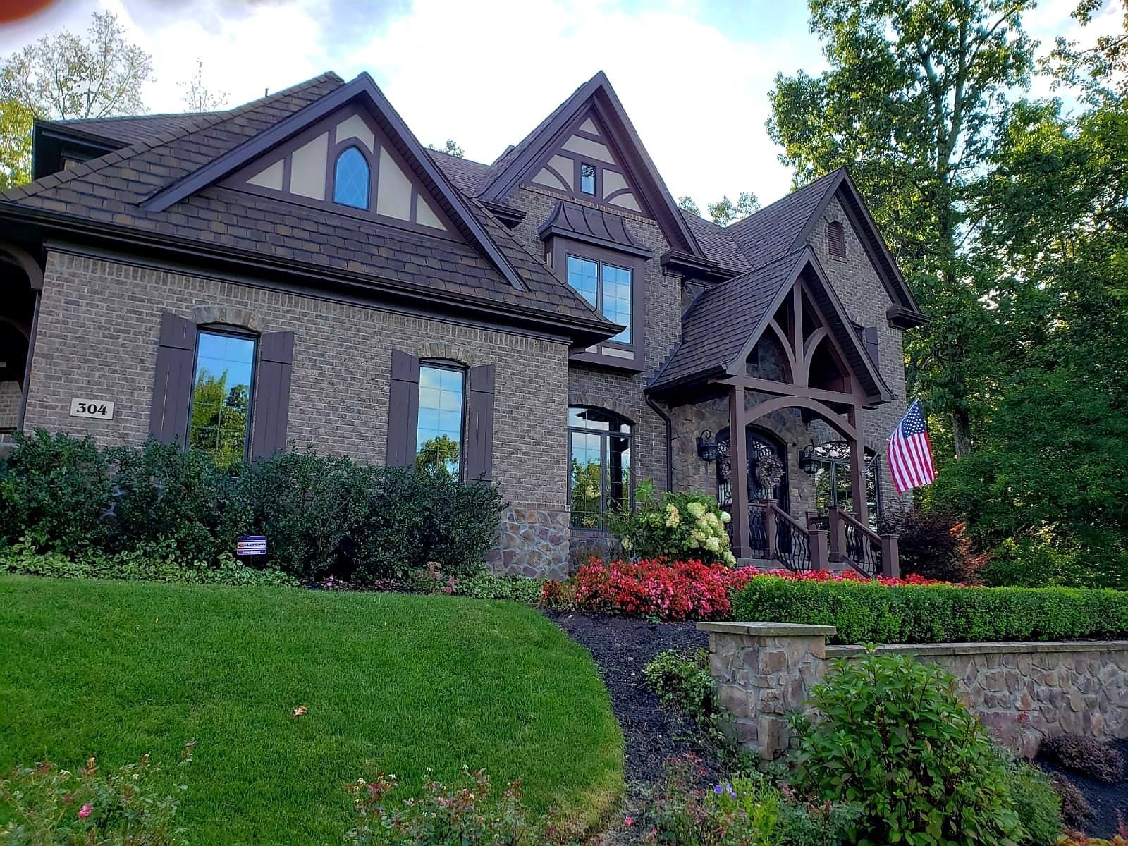 A brick house with a brown roof and trim, a covered porch with an American flag, and a landscaped yard.