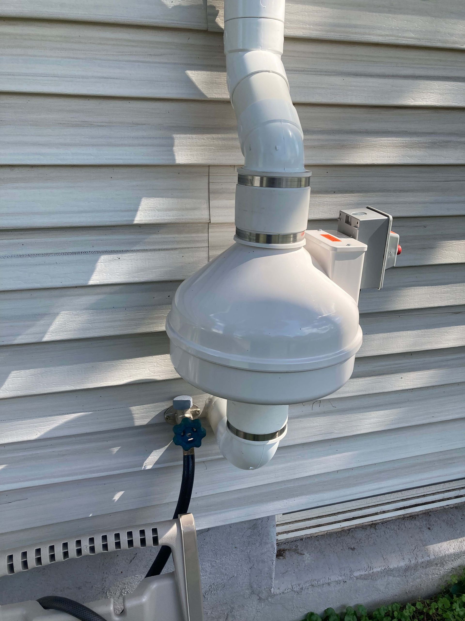 Outdoor wall-mounted exhaust vent with white ducting and a small blue valve on a house exterior