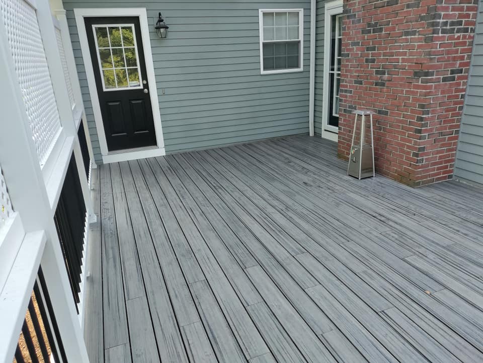 A gray deck with a black door and a brick wall in the background.