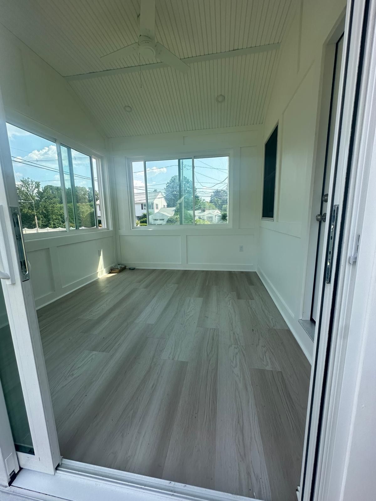 Bright white sunroom with gray wood-look flooring, windows, and sliding glass door; empty space.