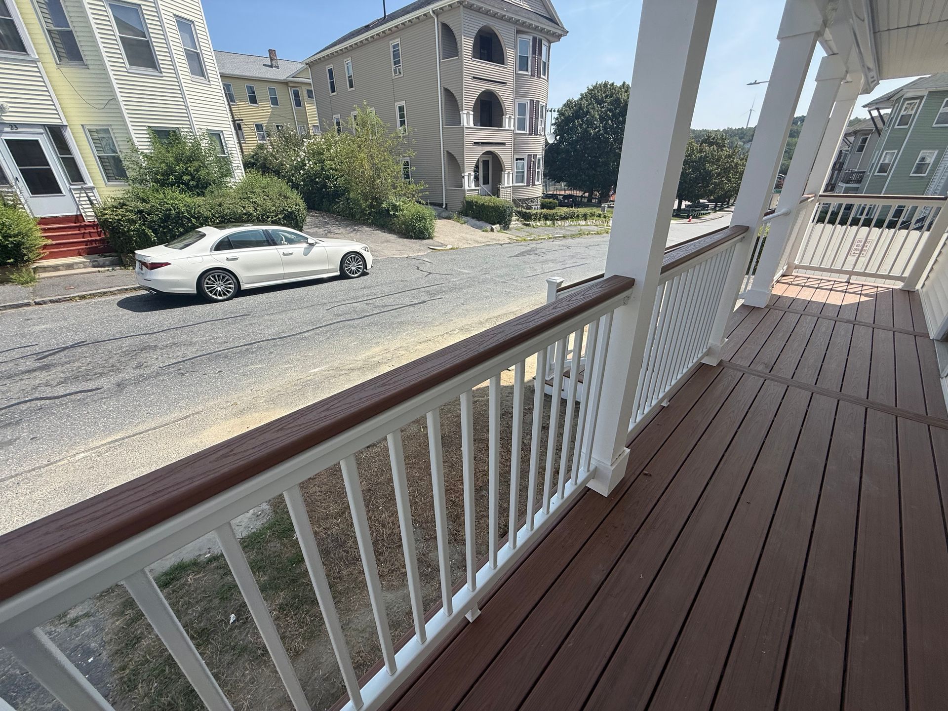 A porch with brown decking and white railing overlooking a street with a white car, and houses.
