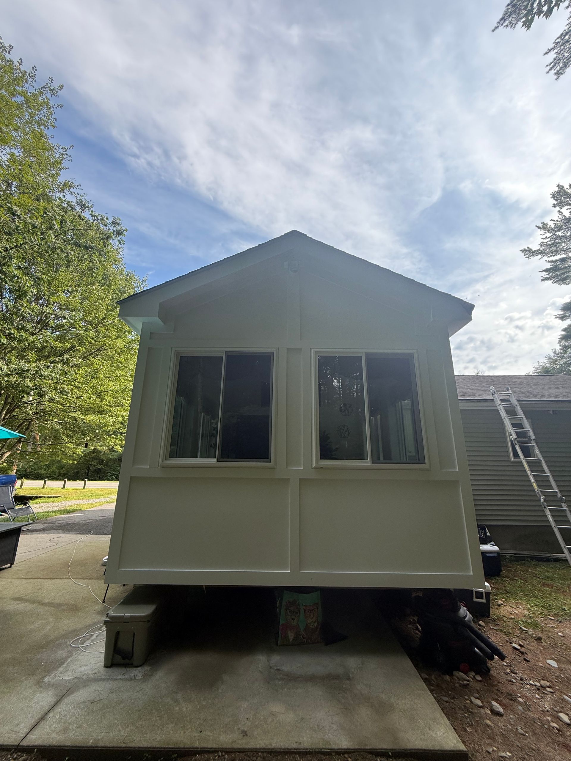 A small white sunroom with two windows, on a concrete base, with trees and a cloudy sky in the background.