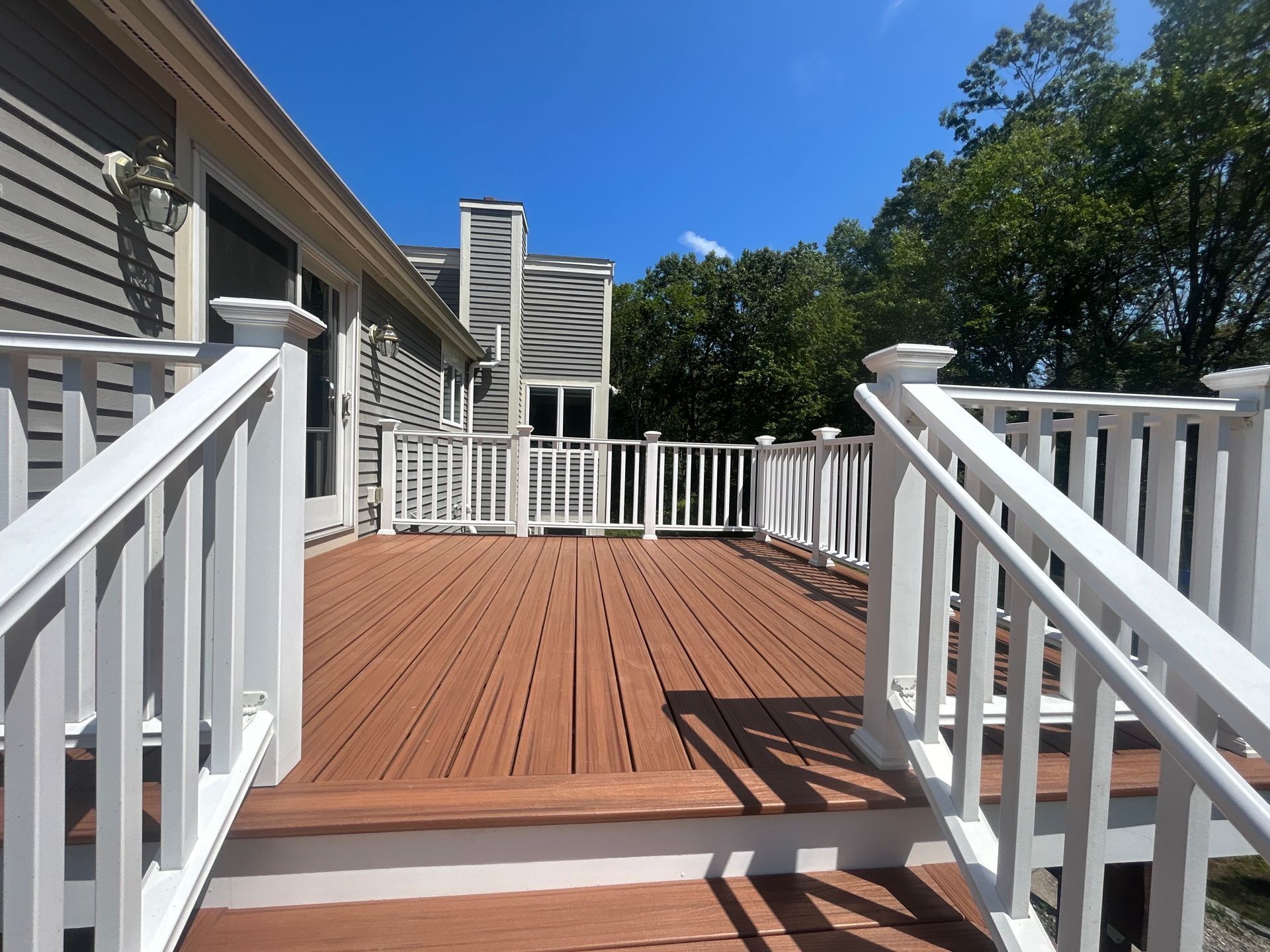 Wooden deck with white railings and brown decking, attached to a grey house under a blue sky.