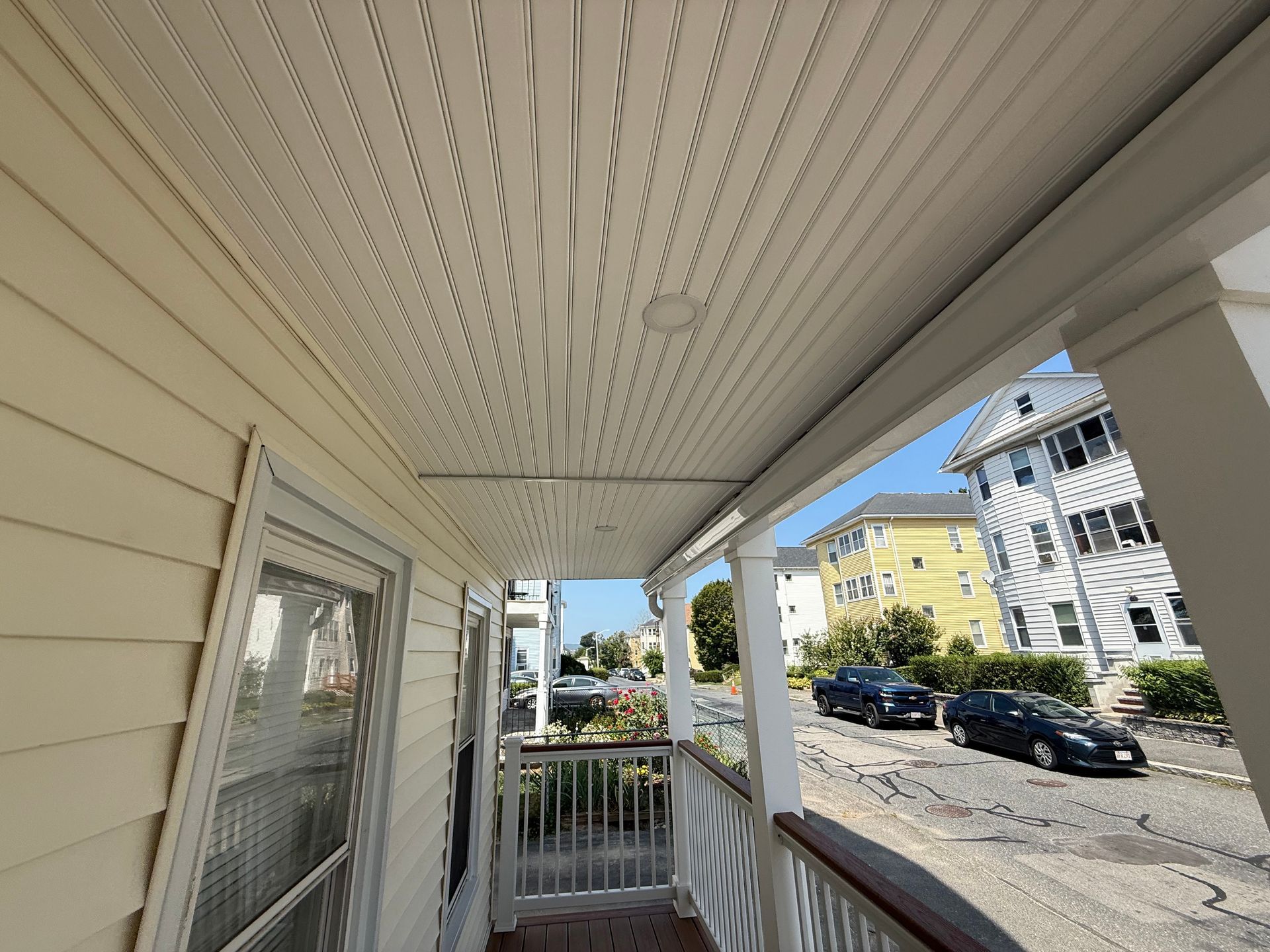 Porch roof with white siding, a doorway, and street view of a sunny day with cars parked.